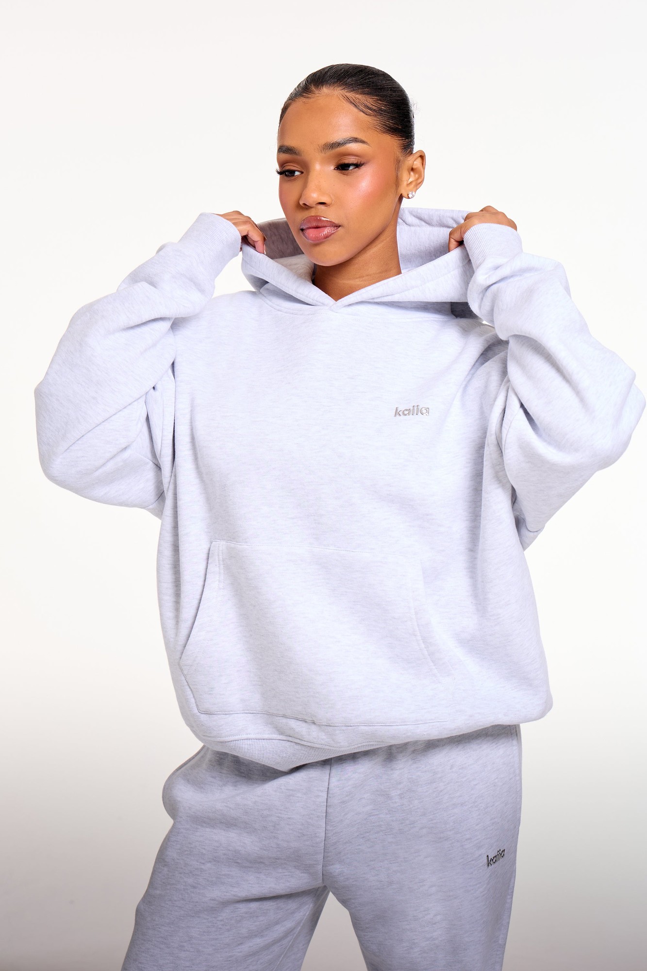 Oversized grey melange hoodie featuring a subtle chrome logo, made from soft material with a front pocket and a spacious hood, styled with matching grey bottoms.