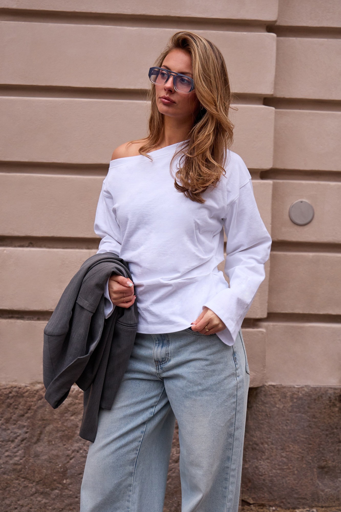 White off-the-shoulder top with a cinched waist, made of soft material, paired with light blue jeans and a gray jacket, styled with glasses and loose waves in hair.