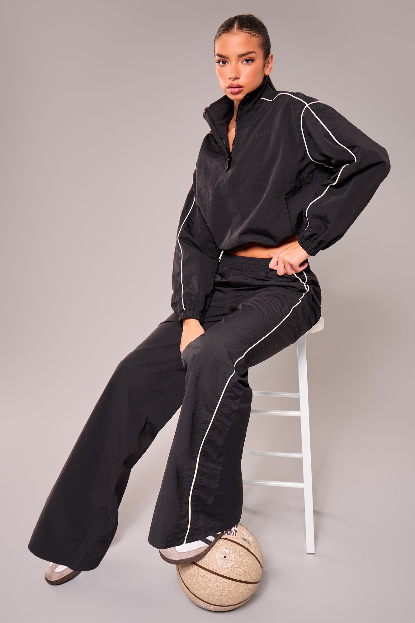 Black wide leg trousers with contrast white piping, made from a lightweight material, featuring a high waist and a relaxed fit, styled with a matching top and sitting on a stool.