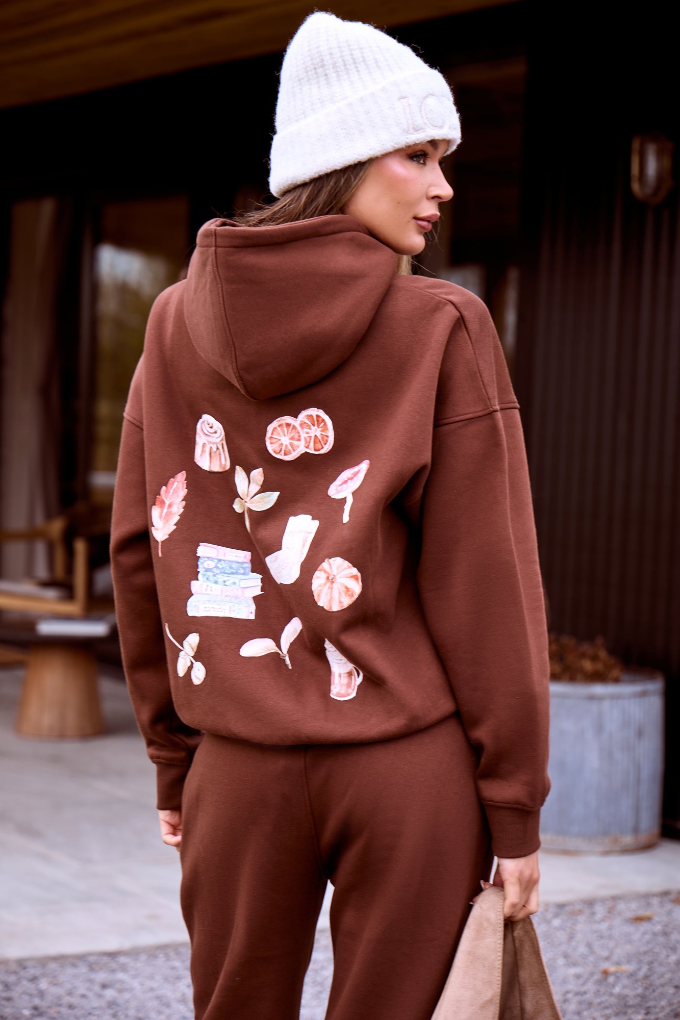 Oversized brown hoodie featuring a cozy graphic design on the back with colorful illustrations, paired with a white knit beanie and matching brown pants.