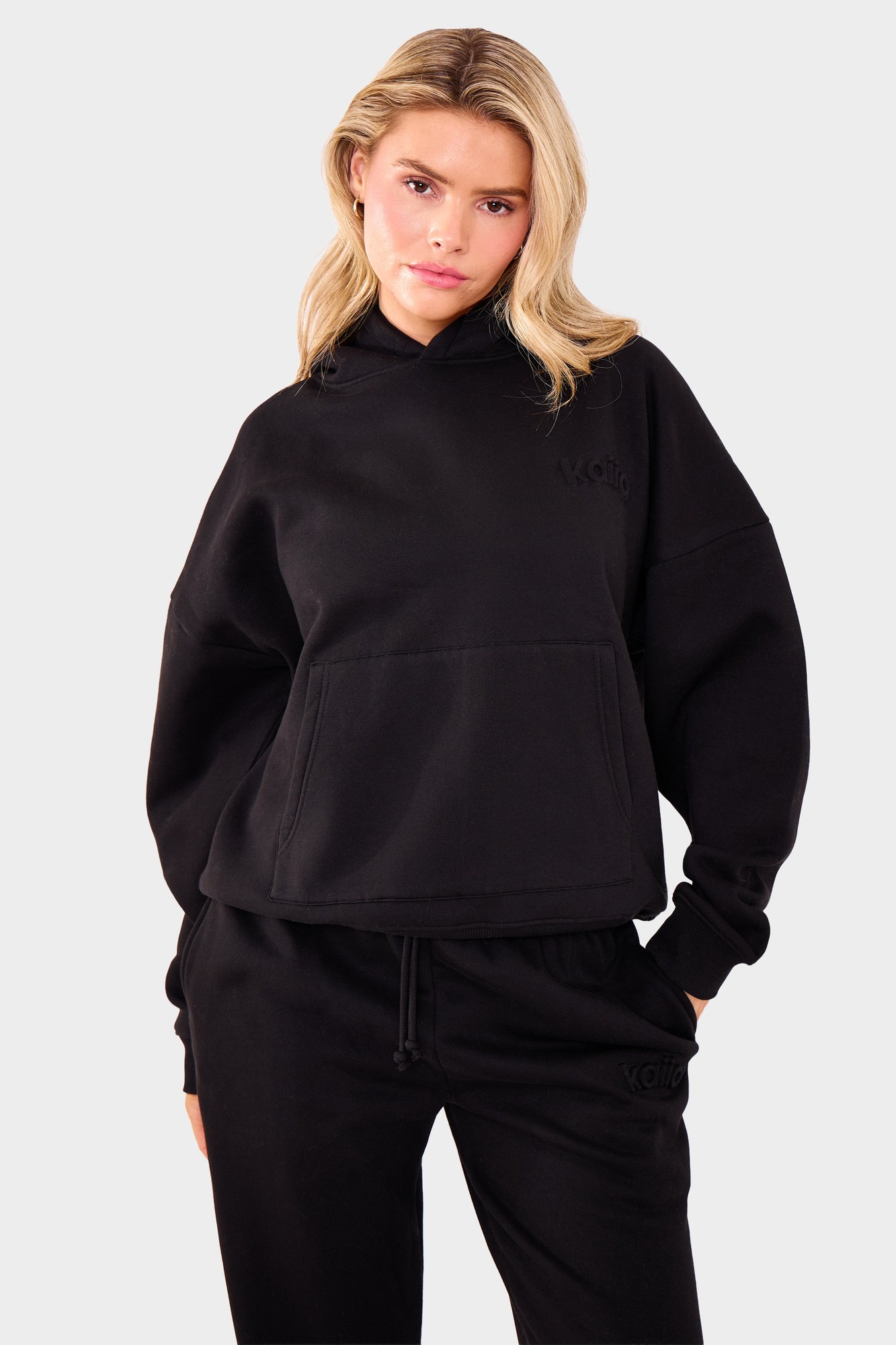 Black oversized hoodie featuring an embossed logo on the chest, made from soft material with a front pocket and a relaxed fit, styled with matching black sweatpants.