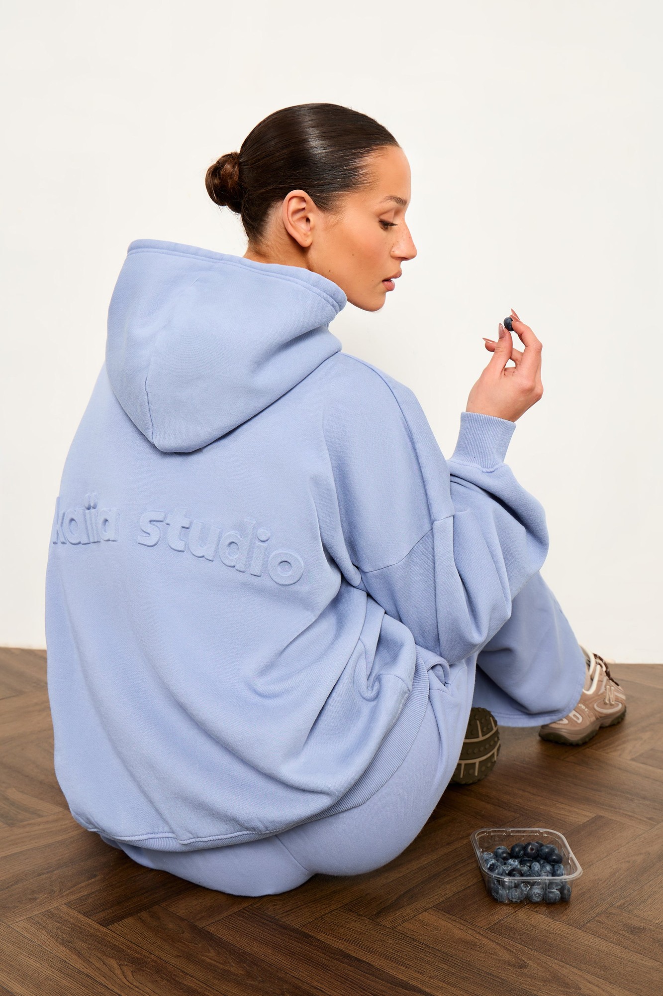 Oversized hoodie in washed periwinkle blue with embossed "kaiia studio" text on the back, made from soft material, styled with matching sweatpants and a casual seated pose.