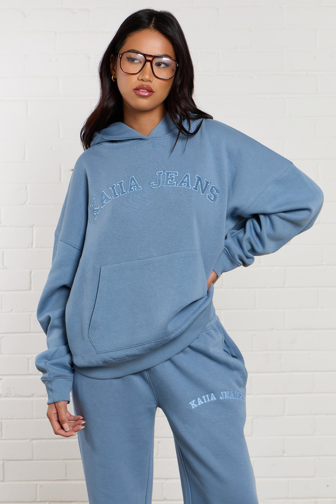 Oversized denim blue hoodie featuring "Kaiia Jeans" applique, a front pocket, and a relaxed fit, styled with matching sweatpants for a casual look.