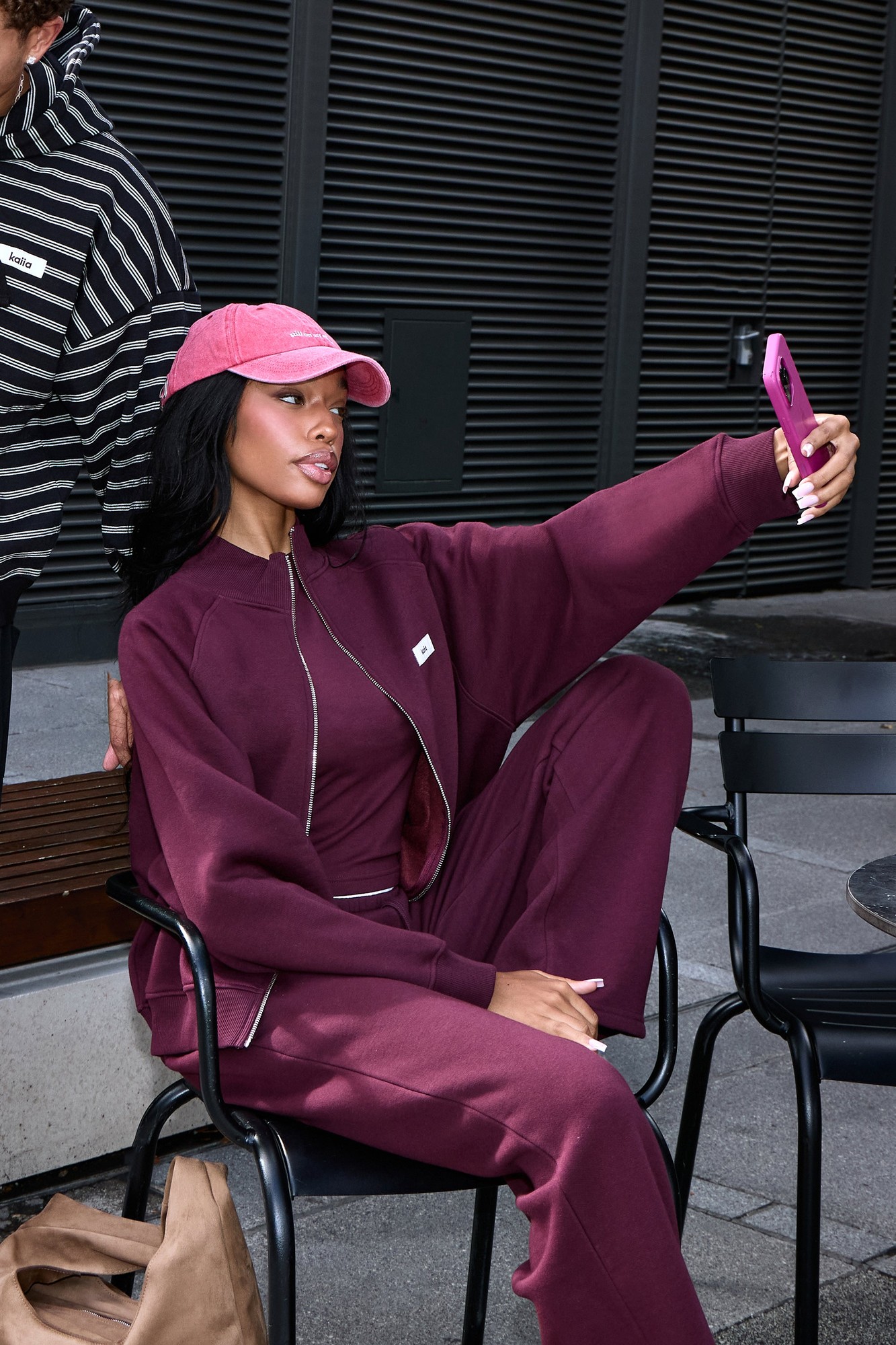 Plum boxy zip-through sweatshirt with a logo, featuring a relaxed fit and ribbed cuffs, worn by a woman sitting on a chair, styled with matching pants and a pink cap.