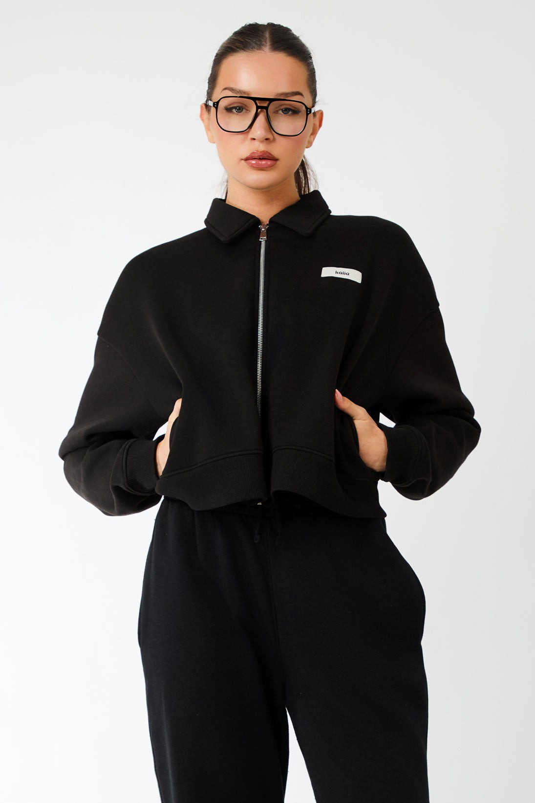 Black collared zip-through sweatshirt by Kaiia, featuring a cropped design, soft material, and a small logo tag on the front, styled with high-waisted black pants.