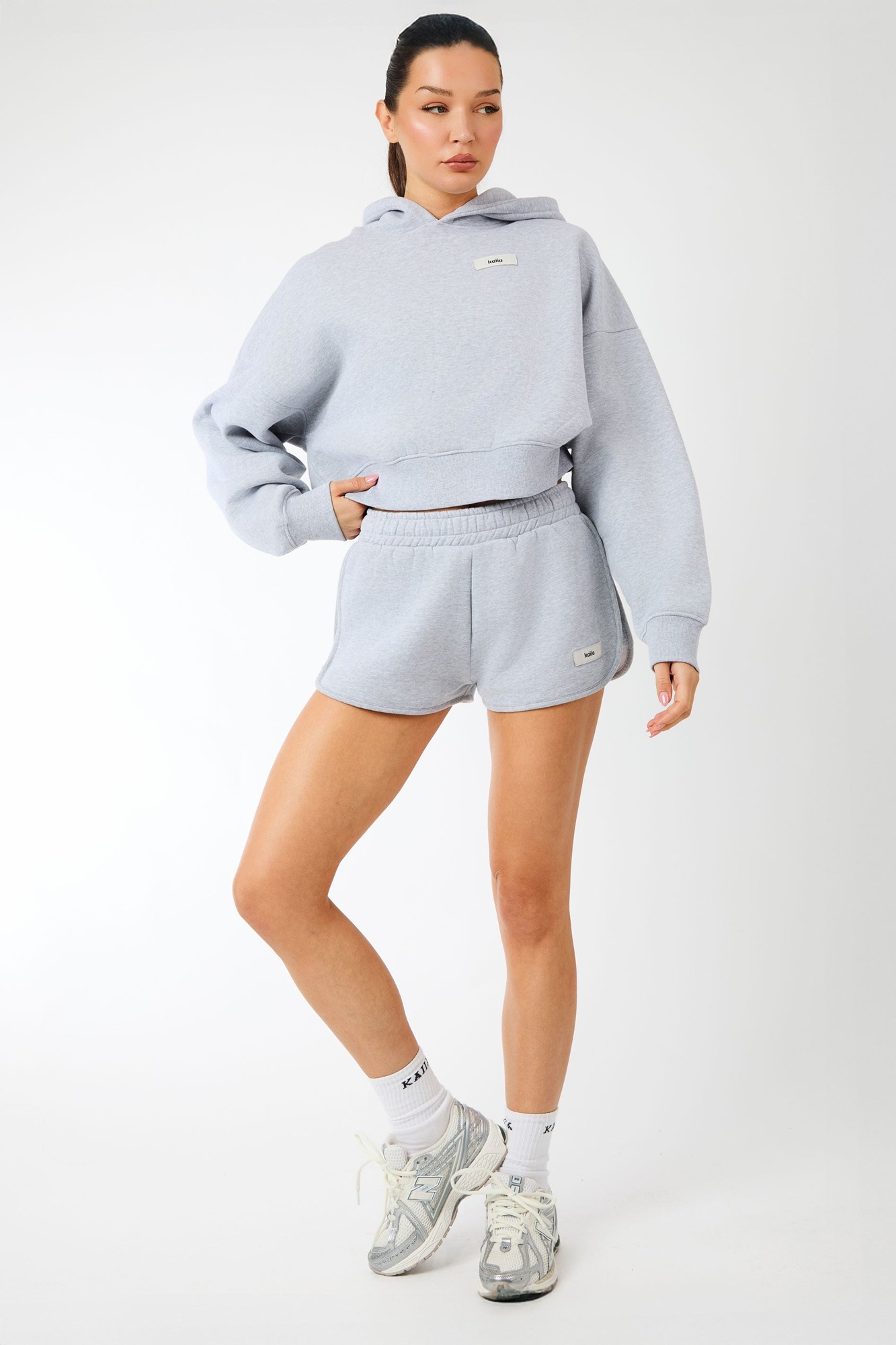 Grey marl mini shorts with a curved hem, featuring an elastic waistband and a small logo tag, styled with a matching cropped hoodie and sneakers.