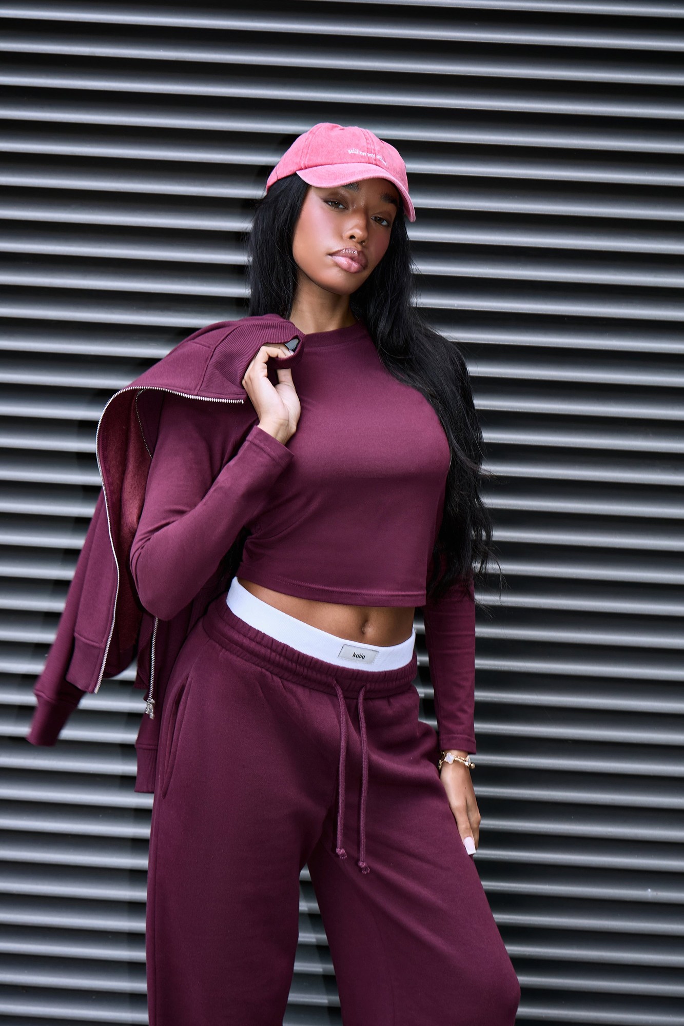 Plum long sleeve baby tee by Kaiia, made from soft material, featuring a fitted design, paired with matching joggers and a pink cap, styled against a textured gray backdrop.