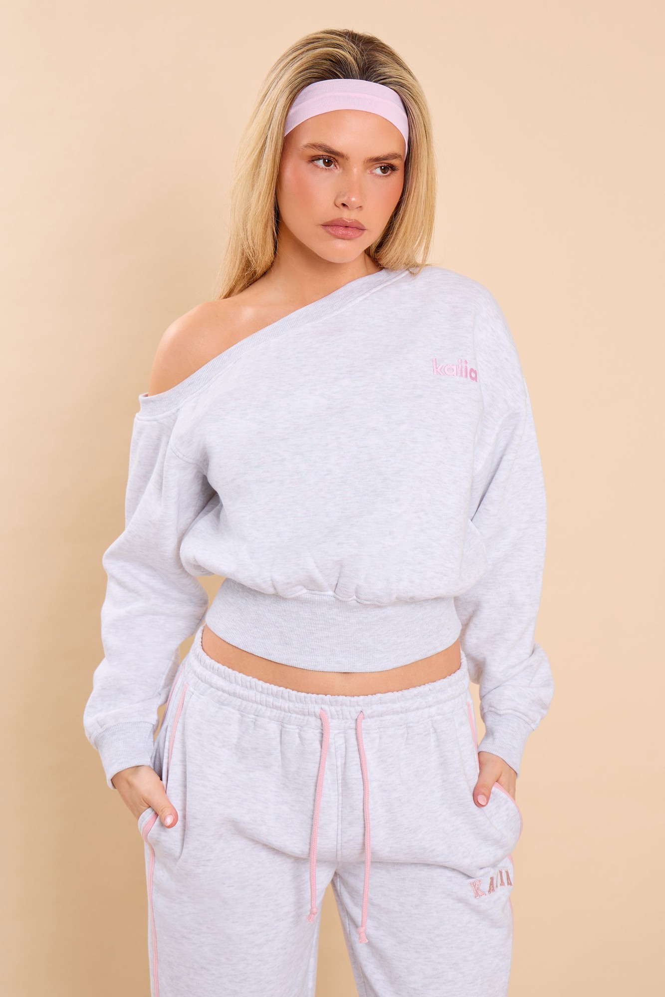 Light grey marl off-the-shoulder sweatshirt with pink "Kaiia" logo, featuring a cropped fit and ribbed hem, paired with matching light grey joggers and a pink headband.