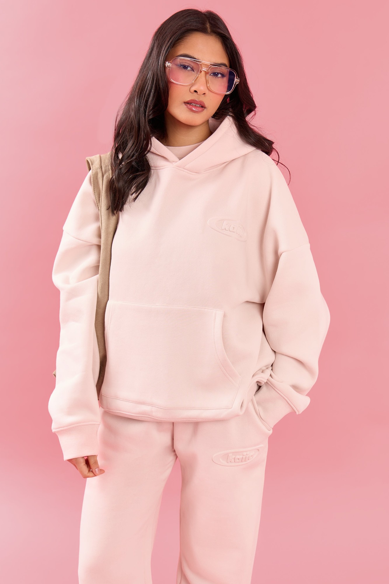 Oversized pink meringue hoodie with an embossed logo, made of soft material, featuring a front pocket and a cozy hood, styled with clear glasses against a pink background.