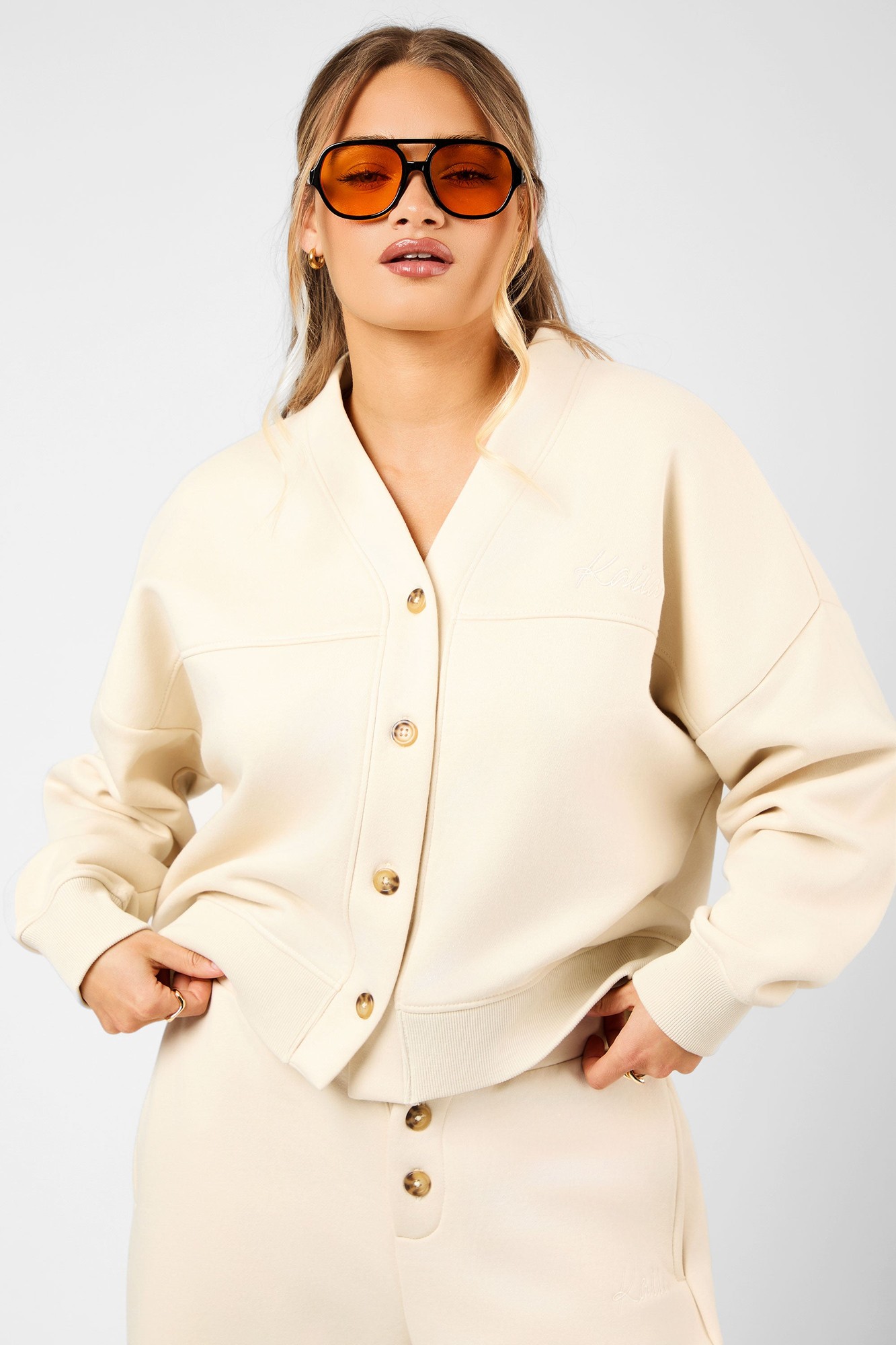 Crème button-down cardigan featuring a soft material, oversized fit, gold buttons, and subtle logo detailing, styled with oversized sunglasses.