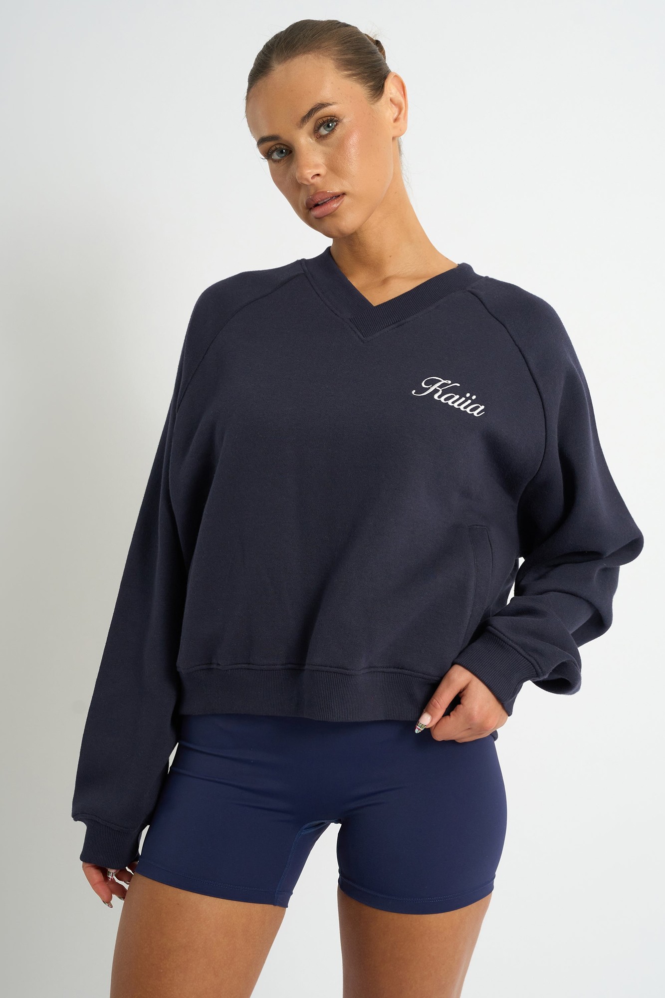 Navy oversized sweatshirt with a V-neck, featuring yellow "Kaiia" script embroidery on the chest, made from a soft material, styled with fitted navy shorts.