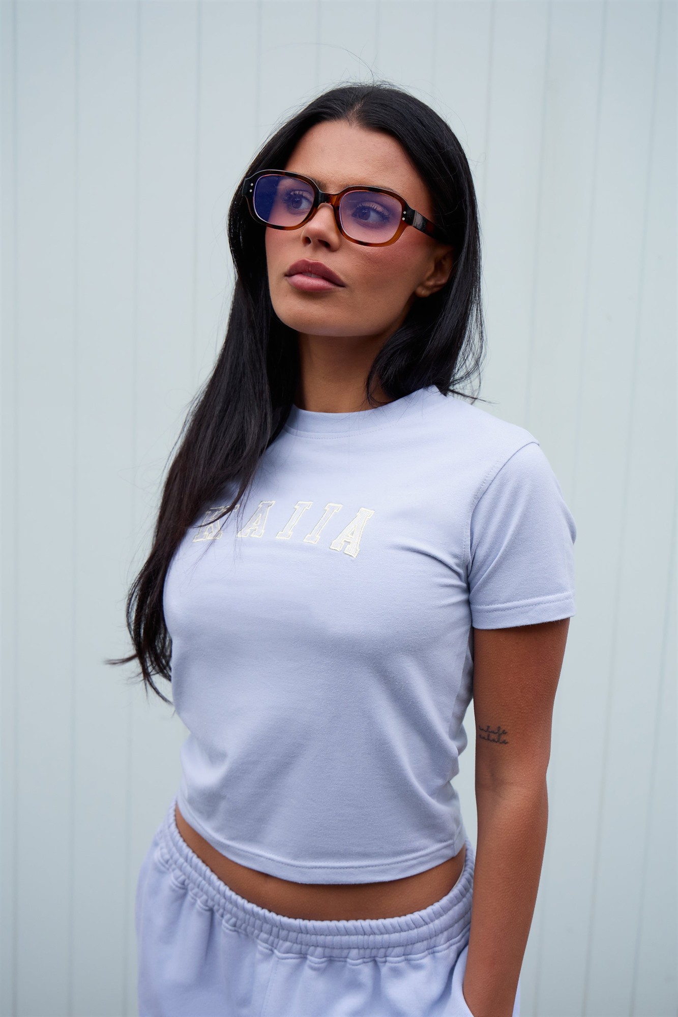 Soft blue baby tee featuring "Kaiia" in white lettering, made from comfortable material, paired with stylish sunglasses and casual light-colored bottoms.