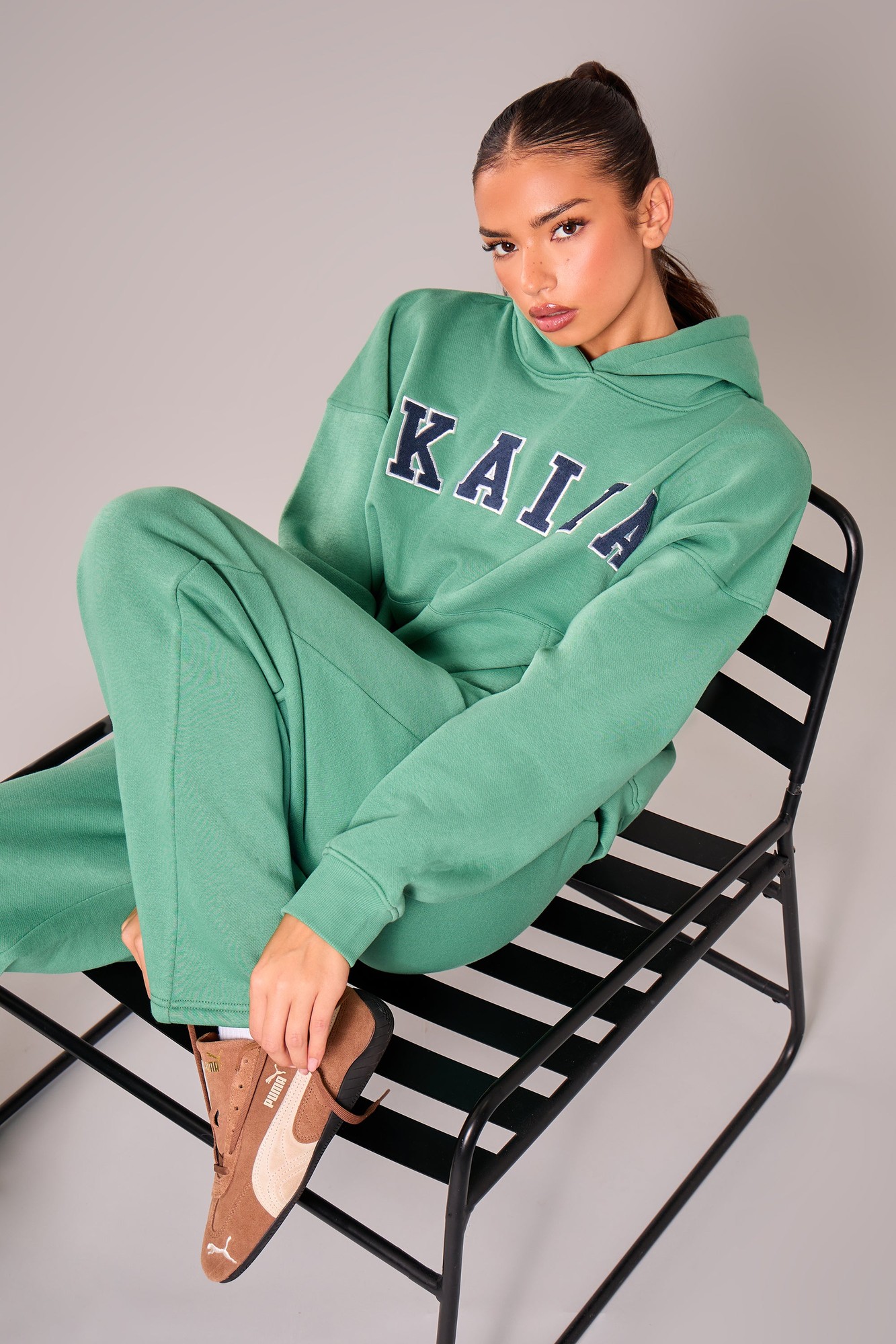 Oversized jade green hoodie with navy "KAIA" slogan, made of soft material, worn with matching pants, seated on a black metal chair.