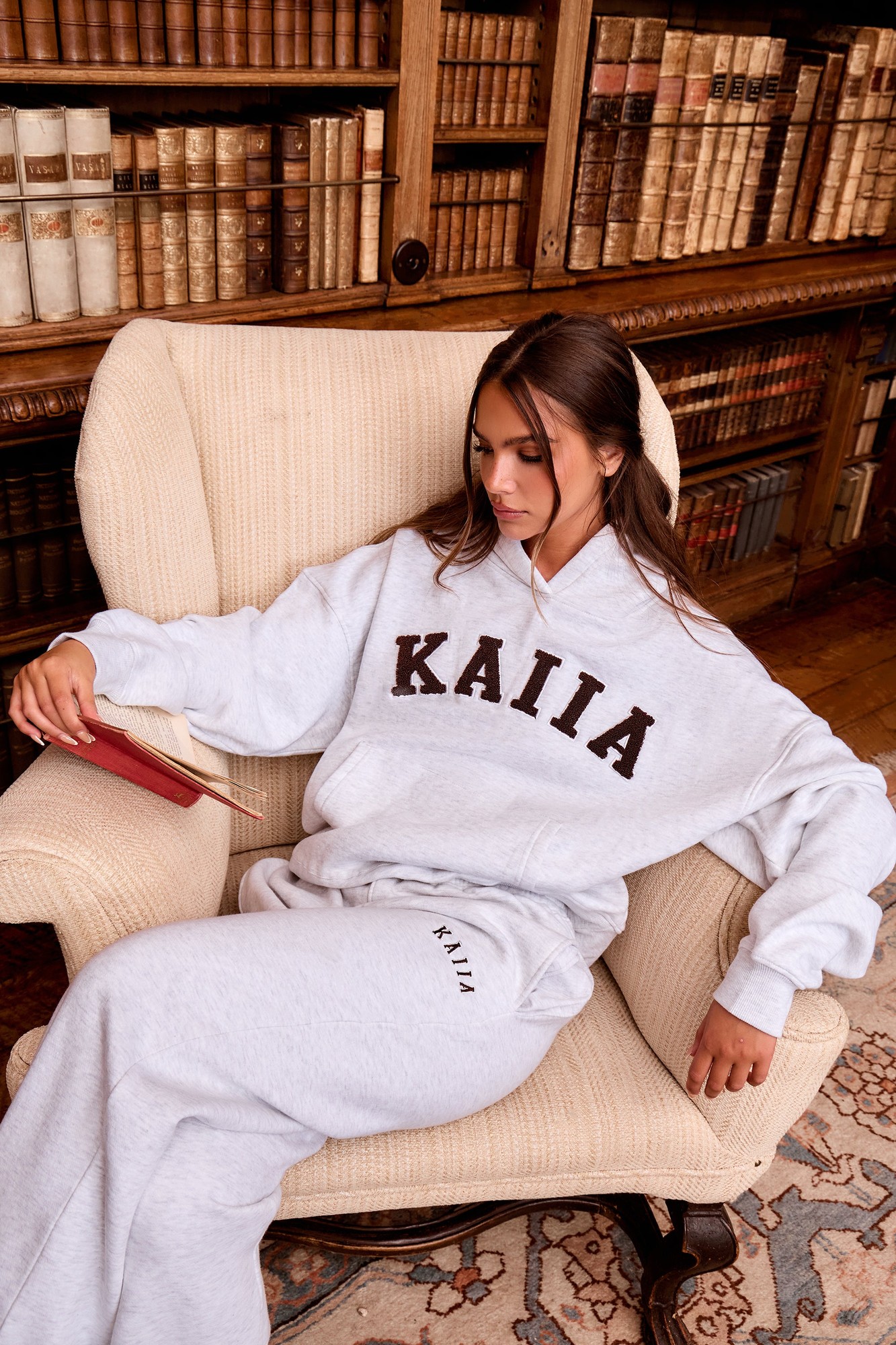 Light grey marl oversized hoodie with dark brown "KAIIA" slogan, paired with matching light grey sweatpants, worn by a seated woman in a cozy library setting.