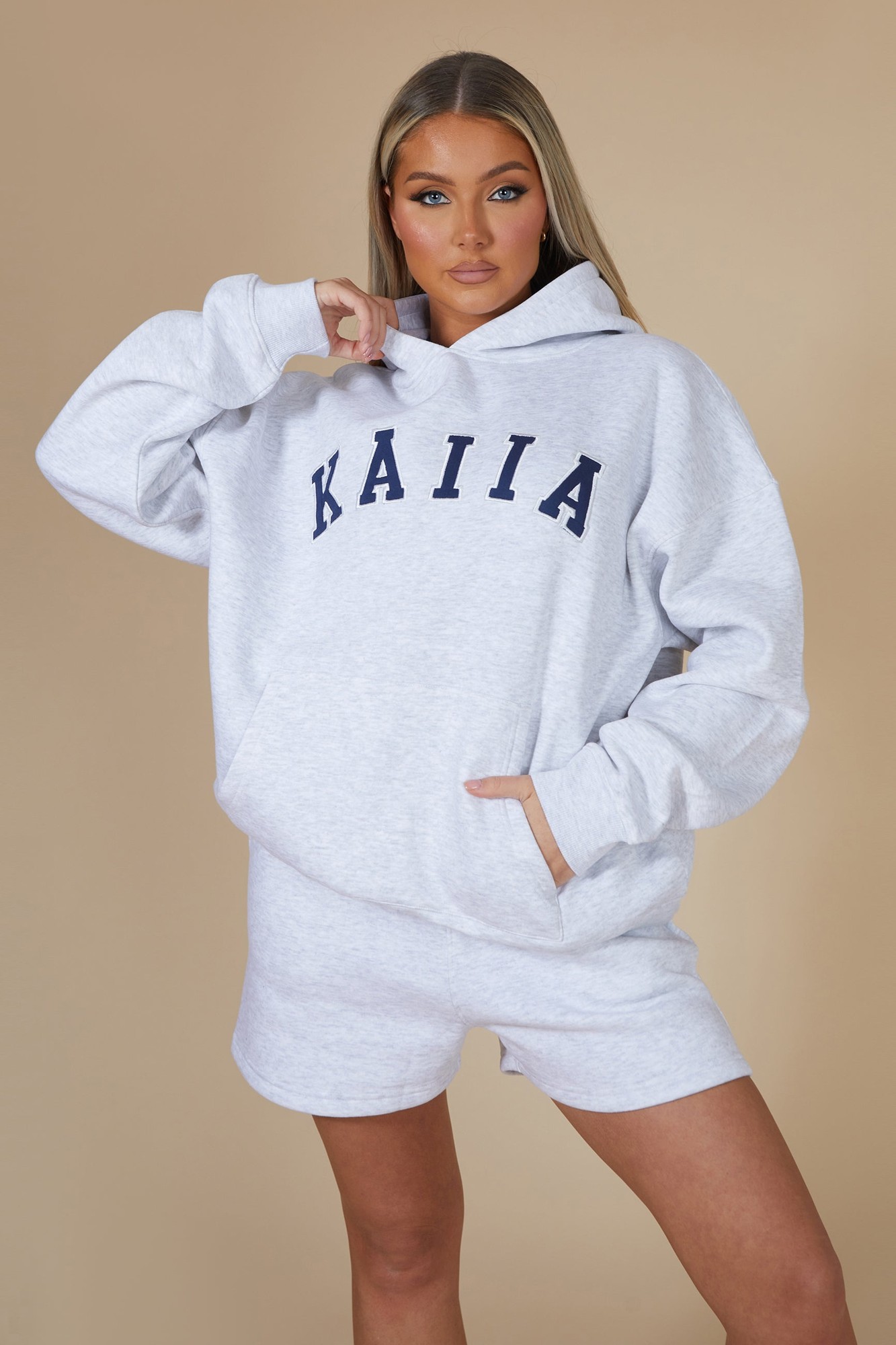 Oversized light grey marl hoodie featuring "KAIIA" in navy lettering, with a front pocket and a cozy hood, styled with matching shorts.
