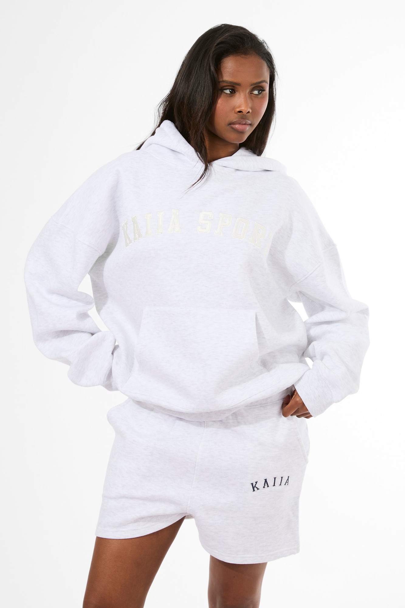 Light grey marl oversized hoodie with cream "KAIJA SPORT" lettering, featuring a spacious hood and relaxed fit, paired with matching light grey shorts.