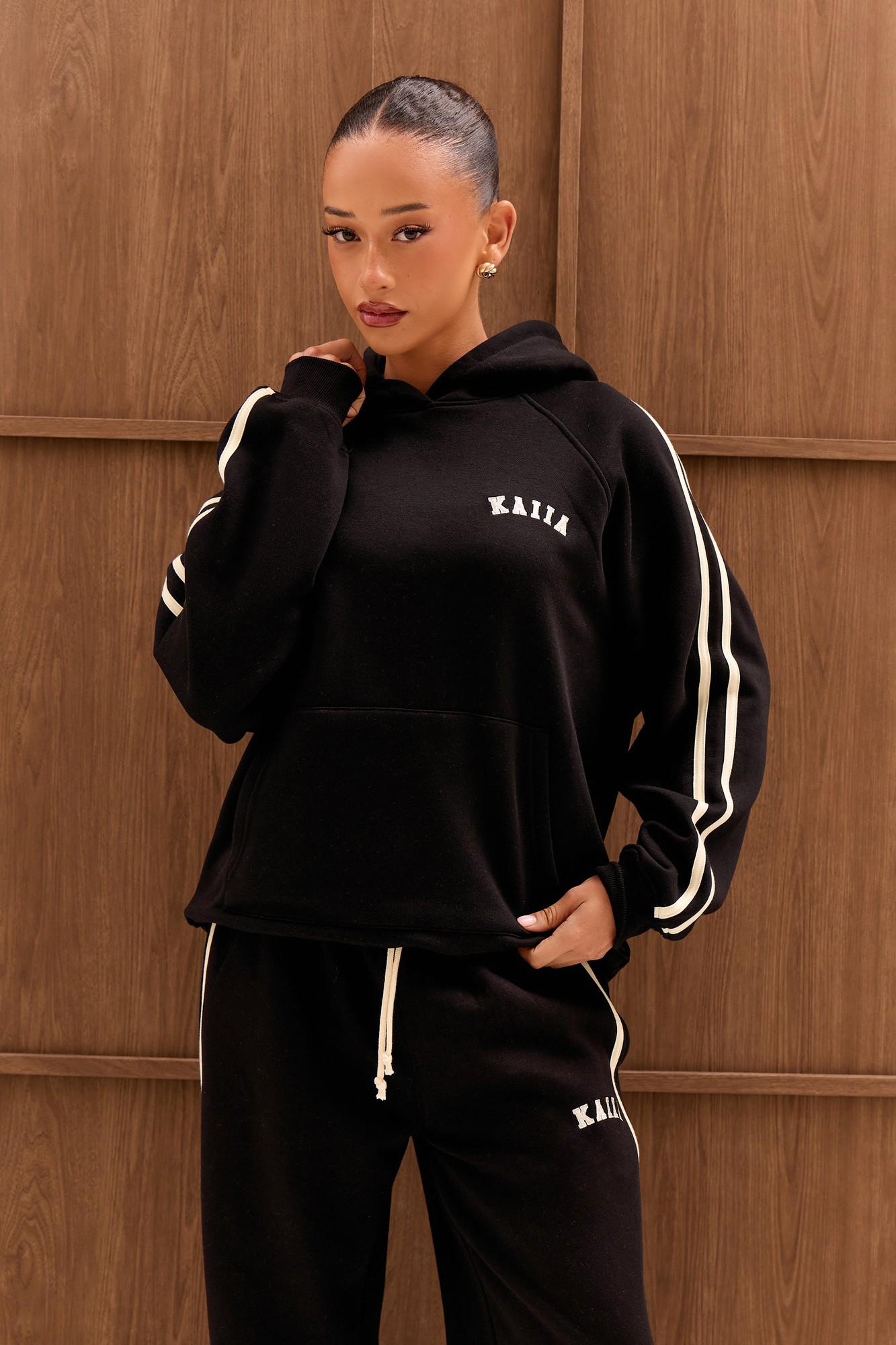 Black oversized hoodie featuring cream side stripes and "KAIIA" logo on the chest, made from soft material, styled with a relaxed fit and front pocket.