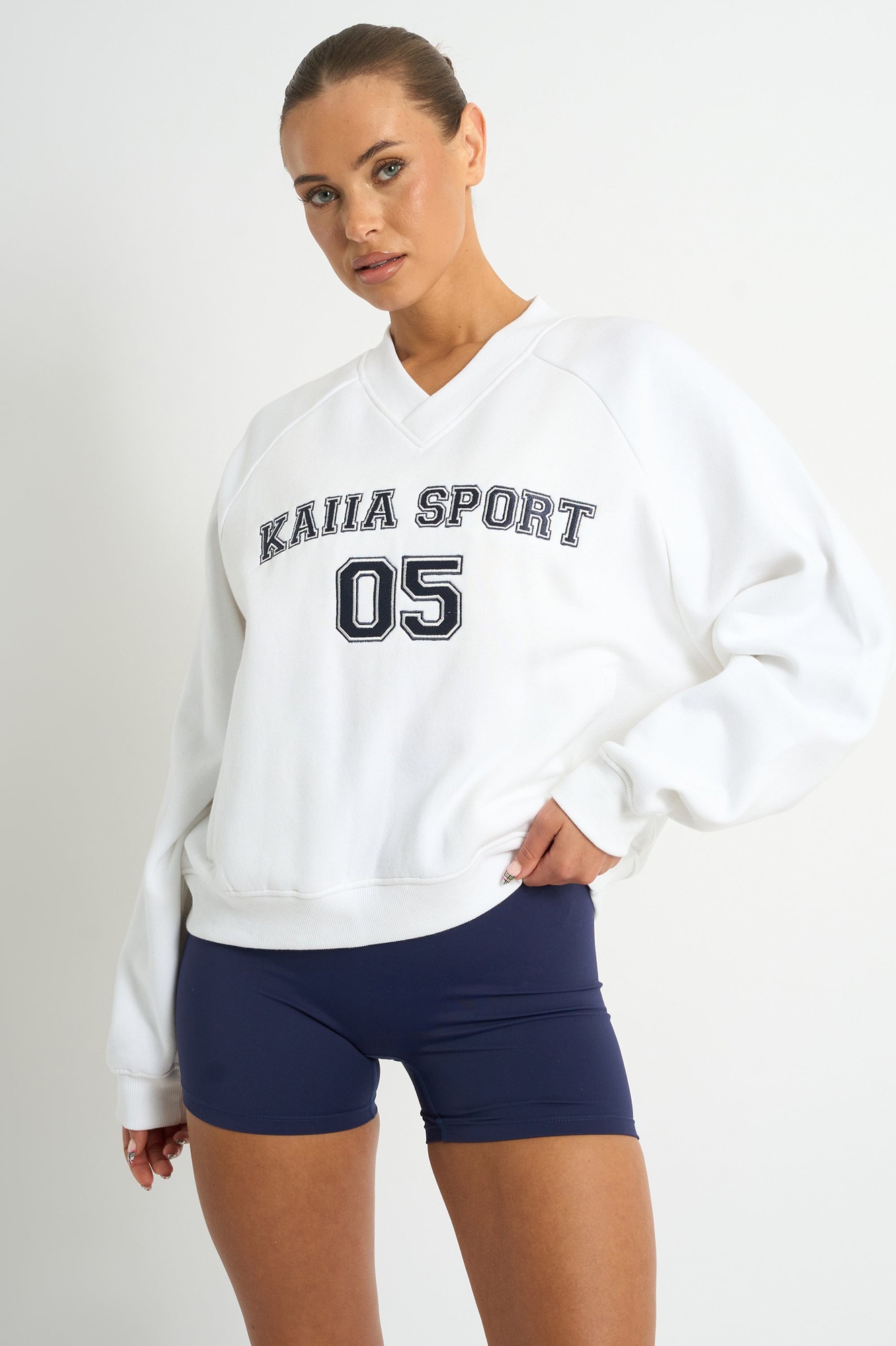 White oversized sweatshirt with navy "KAIIA SPORT 05" lettering, featuring a V-neck and ribbed cuffs, paired with navy shorts.