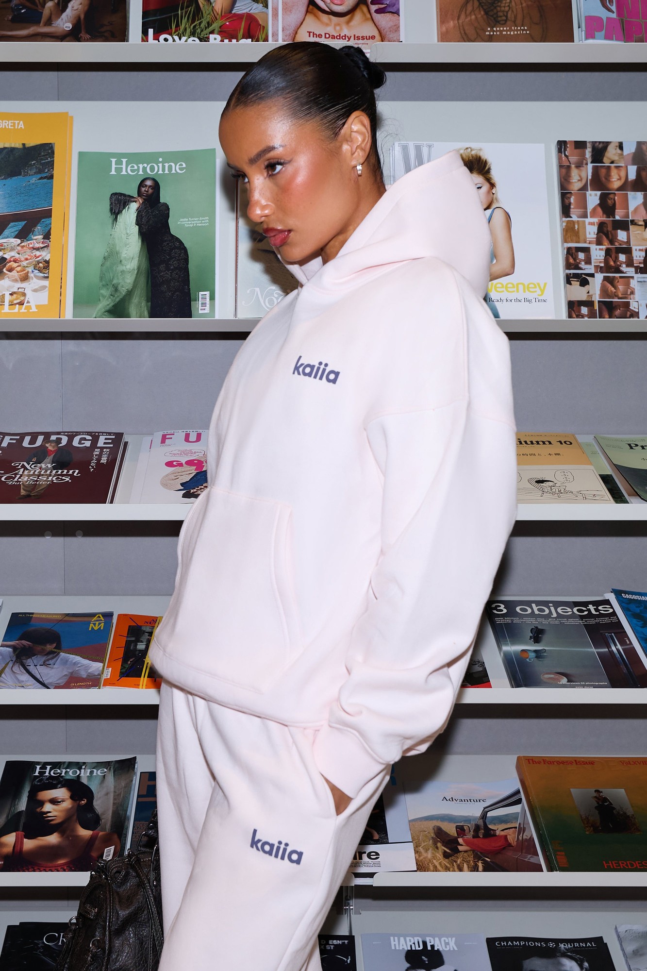 Oversized hoodie in shell pink with dark grey accents, featuring the "kaiia" logo, styled with matching pants and set against a backdrop of magazines.