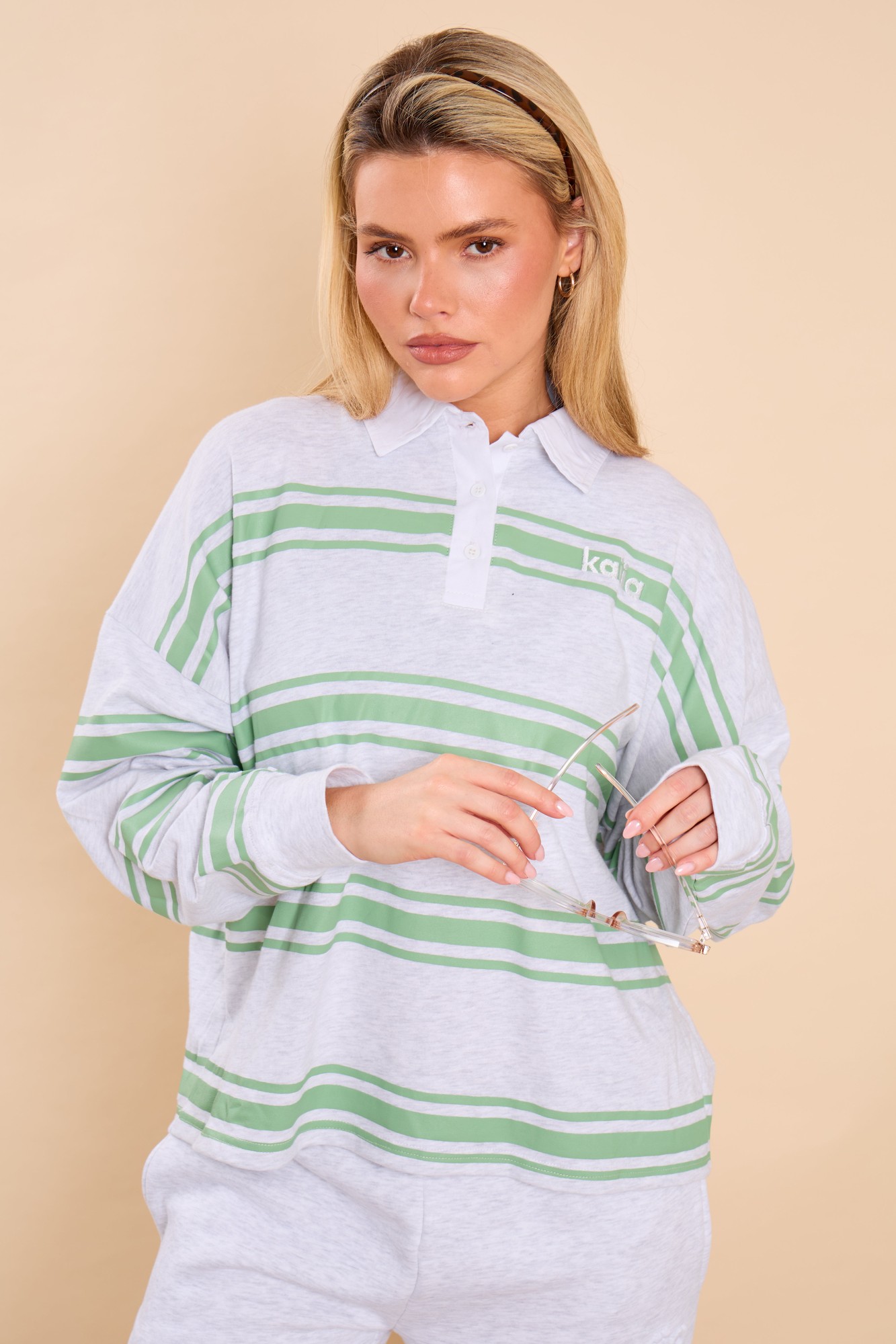 Oversized rugby shirt in light gray with green stripes, featuring a classic collar and the Kaiia logo on the chest, styled with relaxed-fit pants and sunglasses.