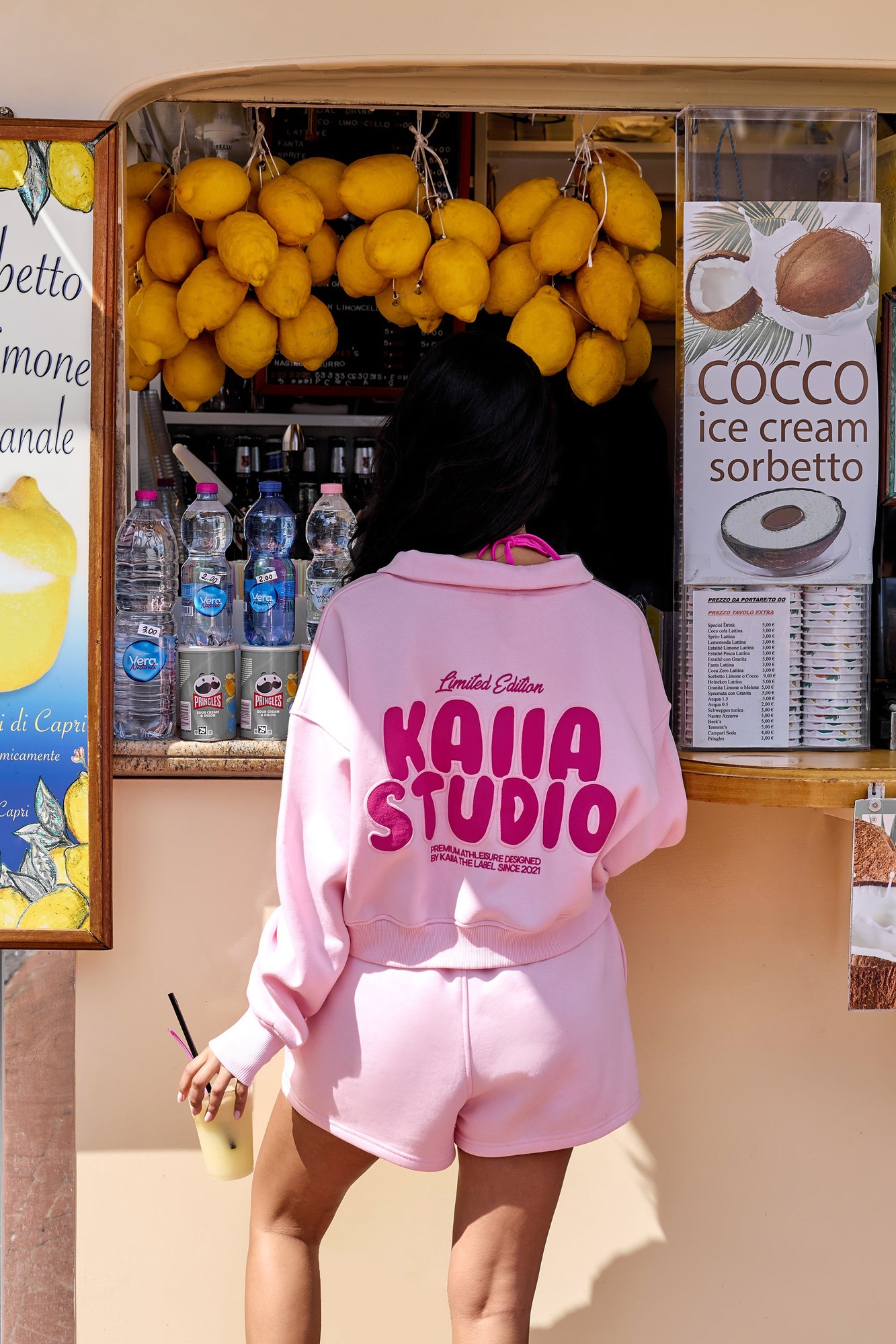 Baby pink quarter zip sweatshirt featuring a bold hot pink "Kaiia Studio" graphic on the back, styled with matching shorts, worn in a vibrant outdoor setting.