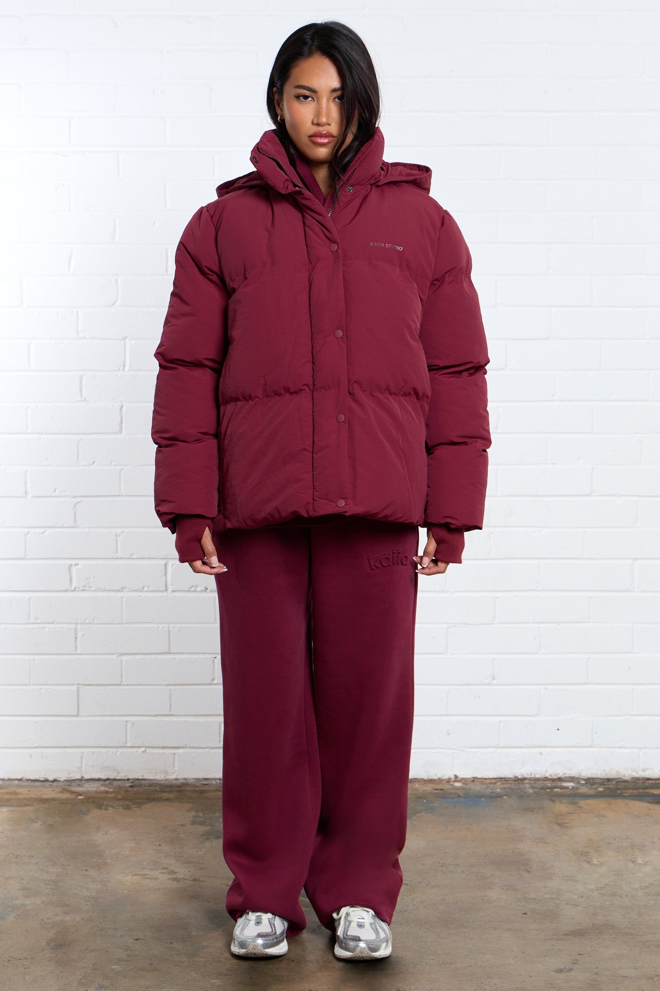Burgundy oversized puffer coat with a hood, featuring a zip and button closure, worn over matching burgundy sweatpants, displayed against a white brick wall.