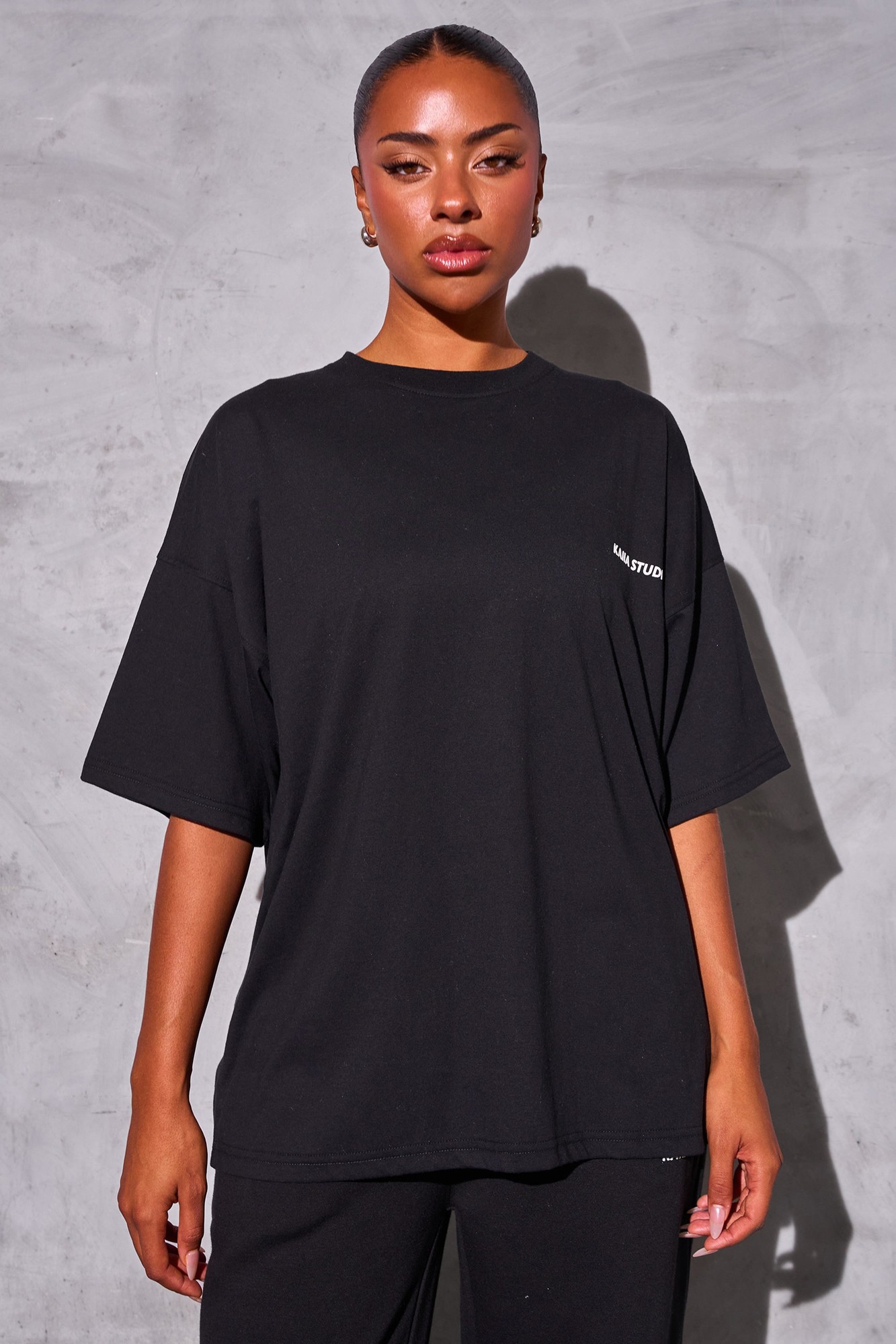 Oversized black tee made from soft cotton featuring a white "Kaiia Studio" logo on the left chest, styled with relaxed sleeves and a comfortable fit.