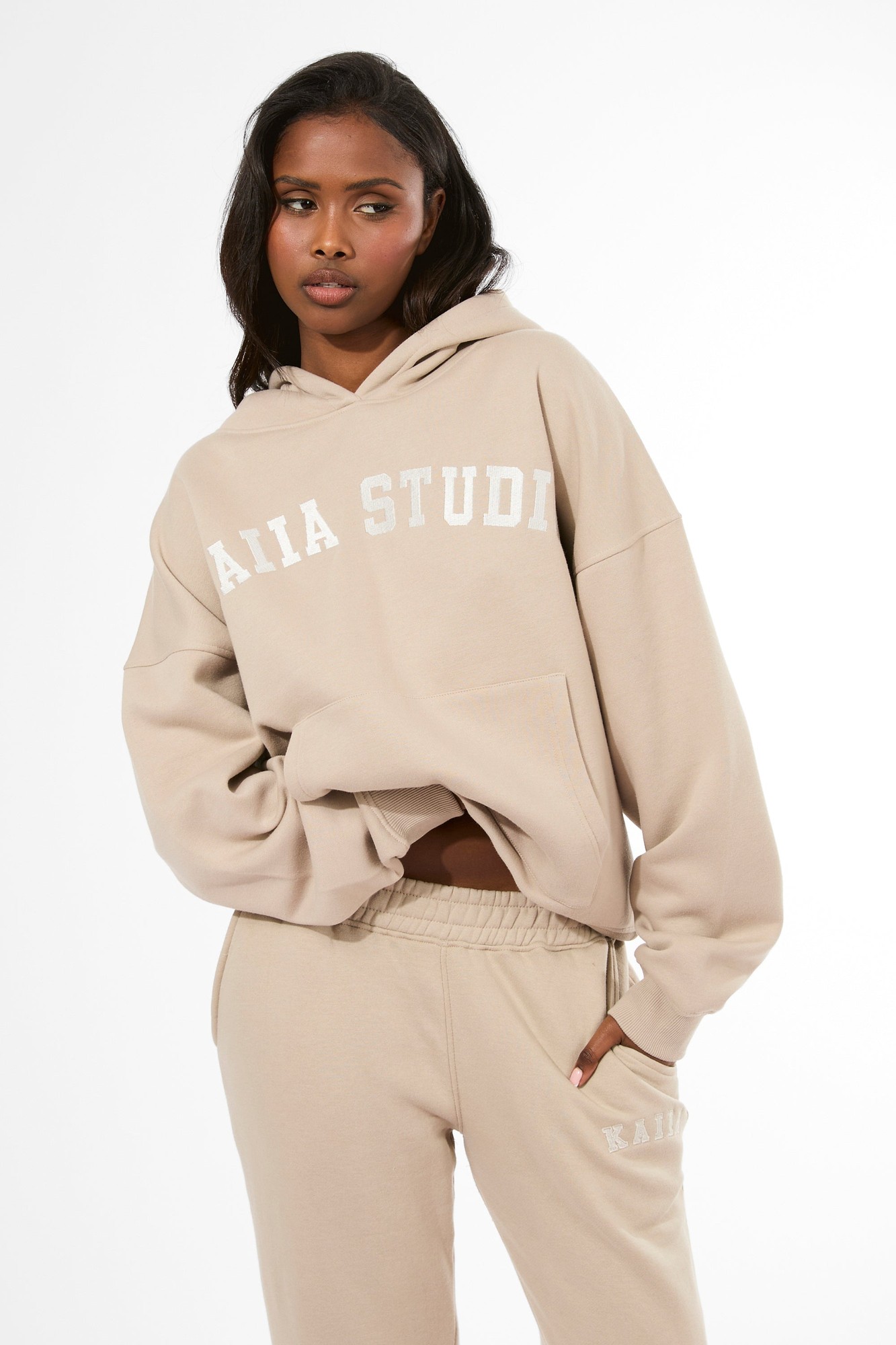 Oversized stone-colored hoodie featuring "KAIIA STUDIO" in white lettering, made of soft material with a front pocket, paired with matching sweatpants.