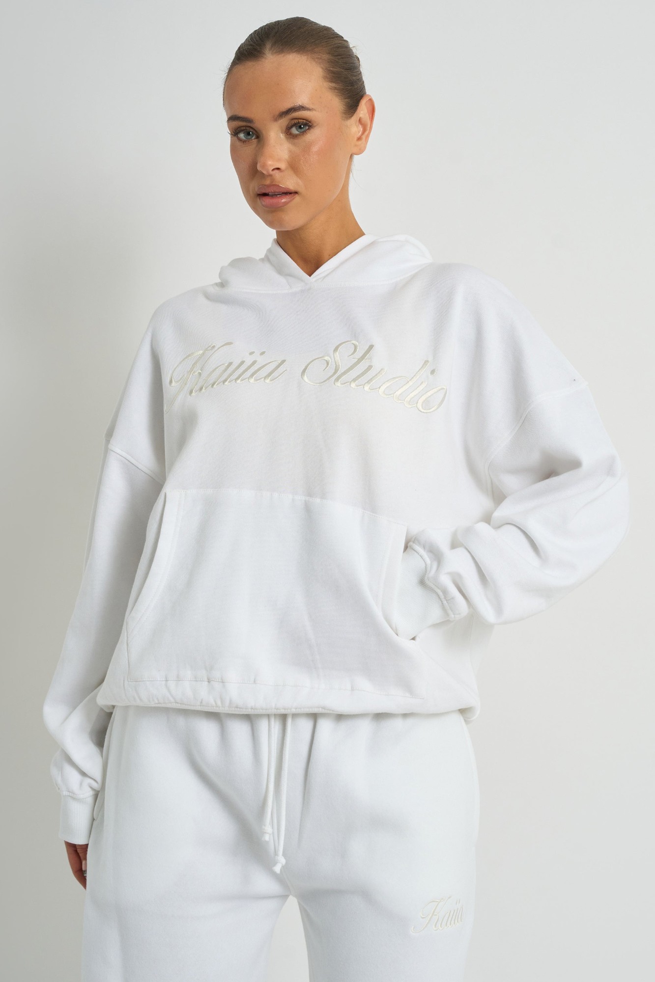 Oversized white hoodie featuring "Kaiia Studio" embroidered in a subtle script, made from soft material with a relaxed fit and a front pocket.