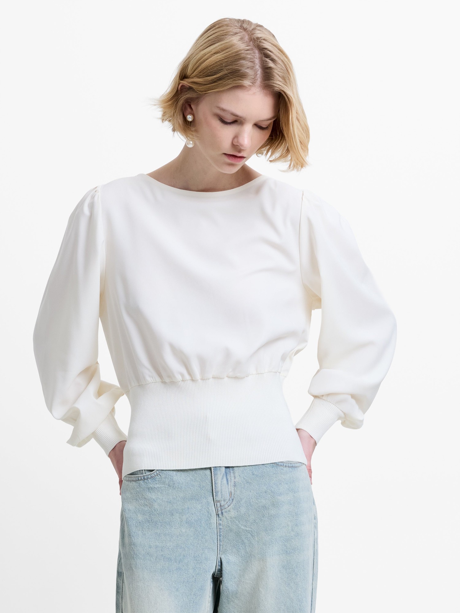 Cream-colored knit jumper with a slash neck, featuring puffed long sleeves and a ribbed waistband, styled with light blue denim jeans.