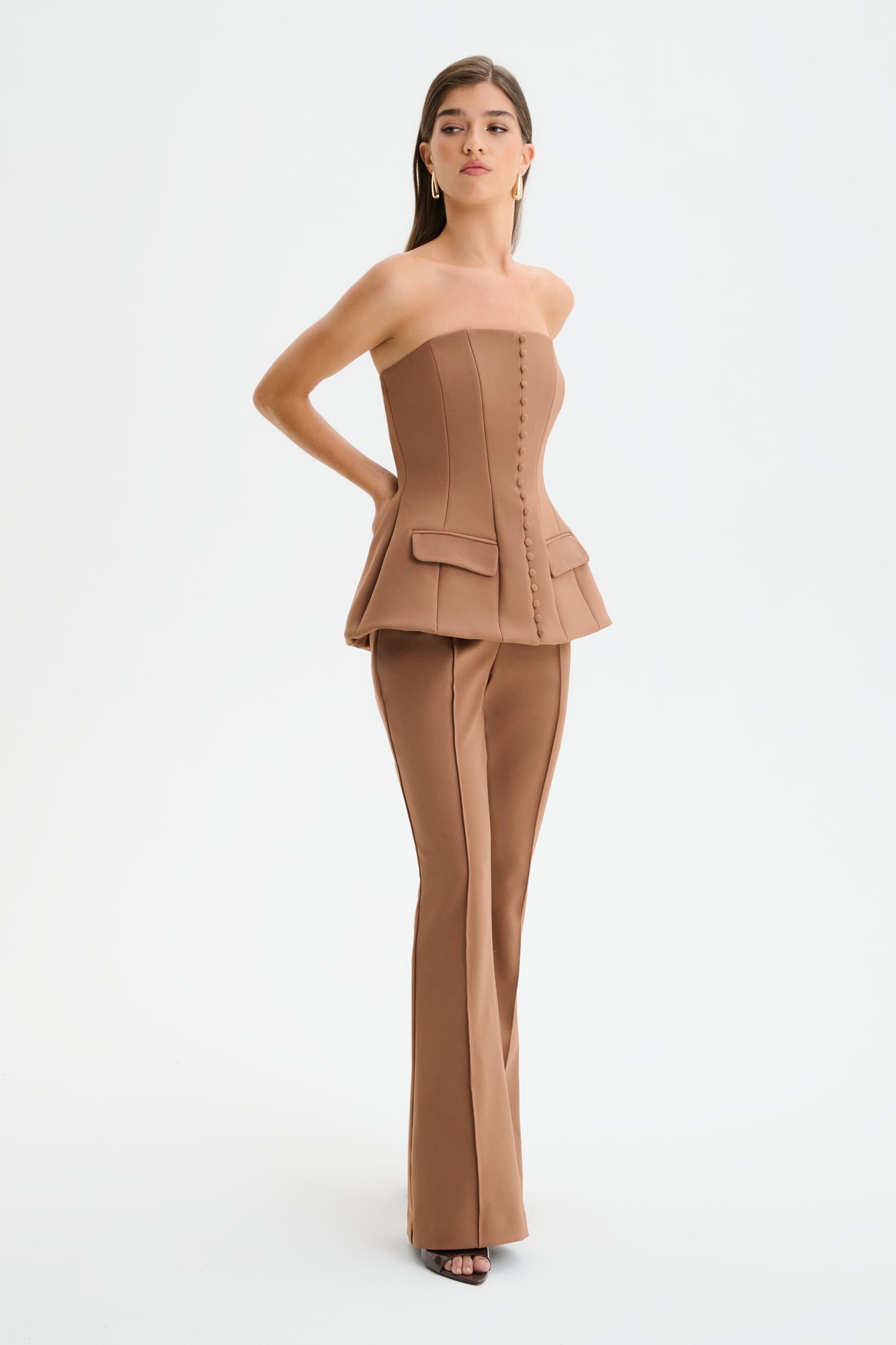 Structured strapless jumpsuit in brown featuring a fitted bodice with button detailing, peplum waist, and flared trousers, made from a smooth, stretchy material.