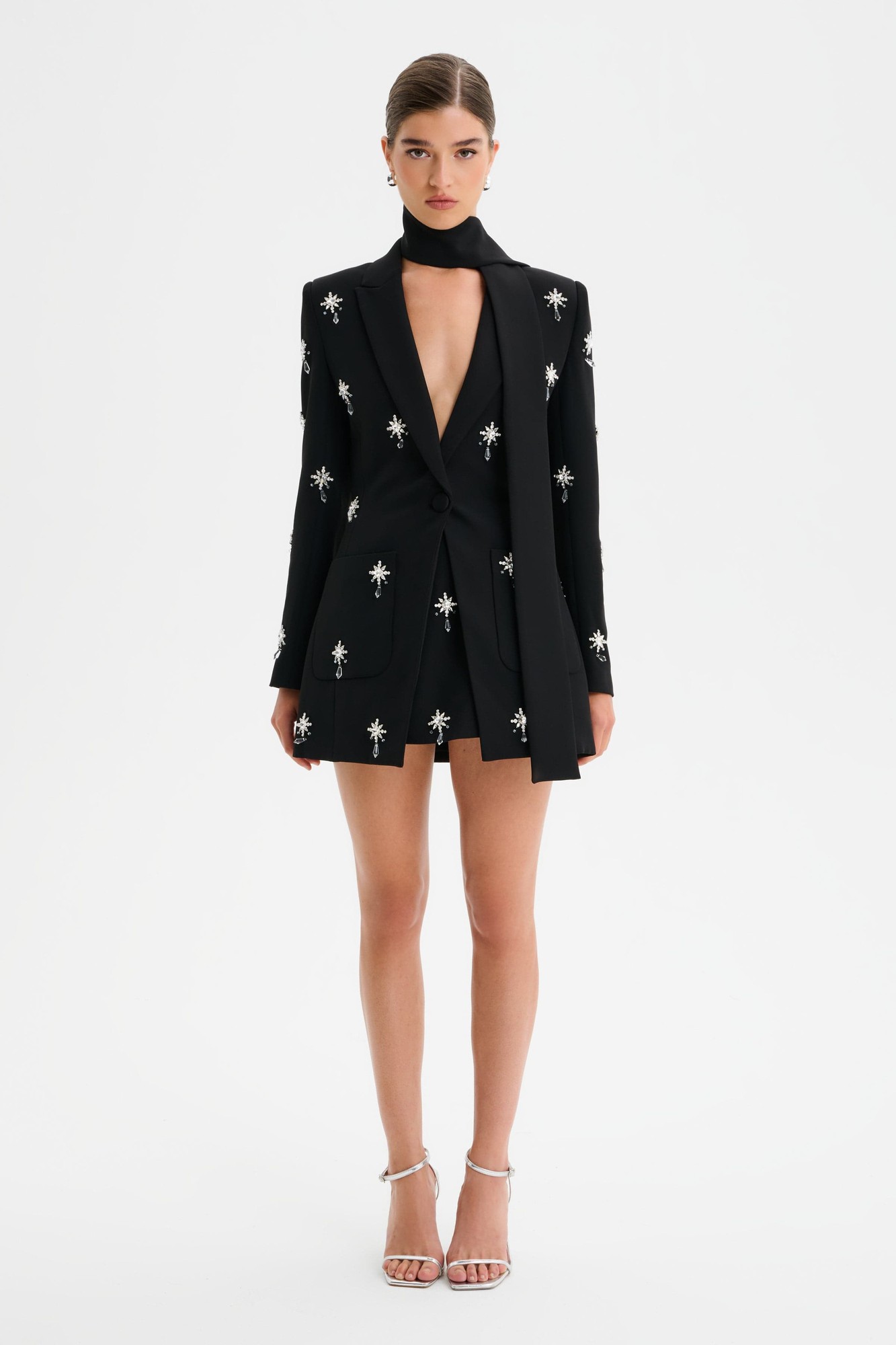 Black tailored jacket with jewel embellishments, deep V-neck, and matching scarf; styled with shorts and strappy heels, showcasing a chic, elegant look.