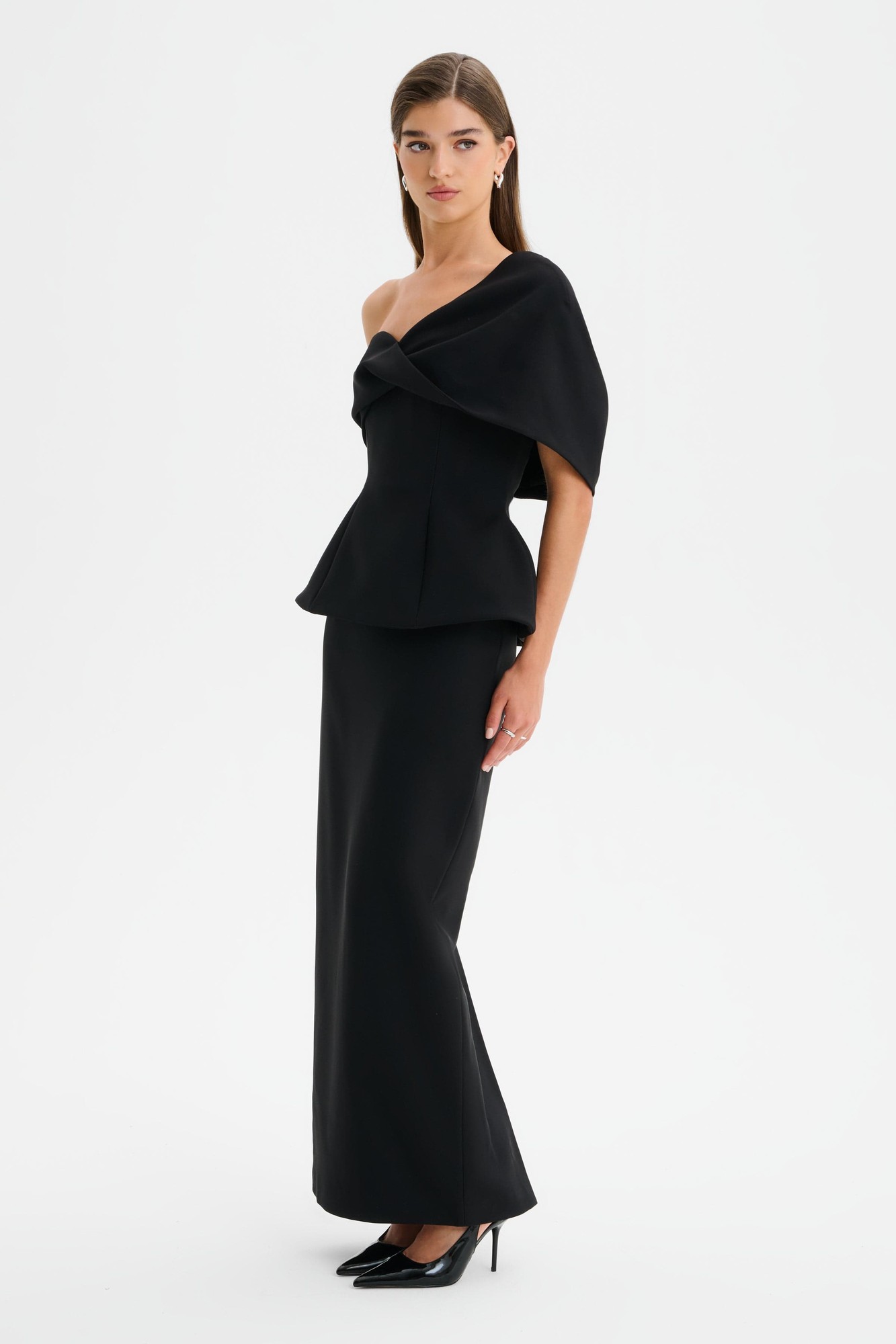 Black maxi dress featuring a twisted cape shoulder design, fitted bodice, and flowing skirt, crafted from a sleek material, styled with pointed black heels.