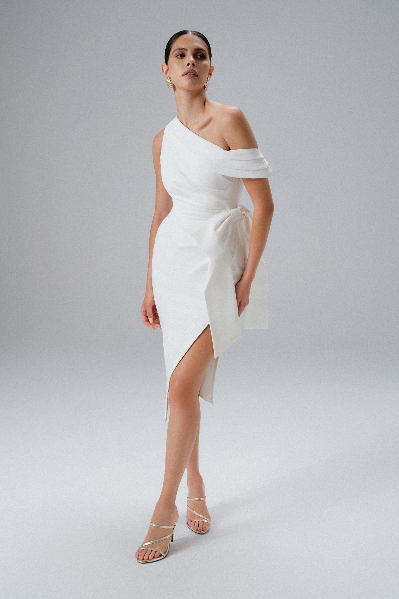 White one-shoulder midi dress featuring pleated detailing and a bow at the waist, made from a smooth material, styled with strappy heels and elegant accessories.