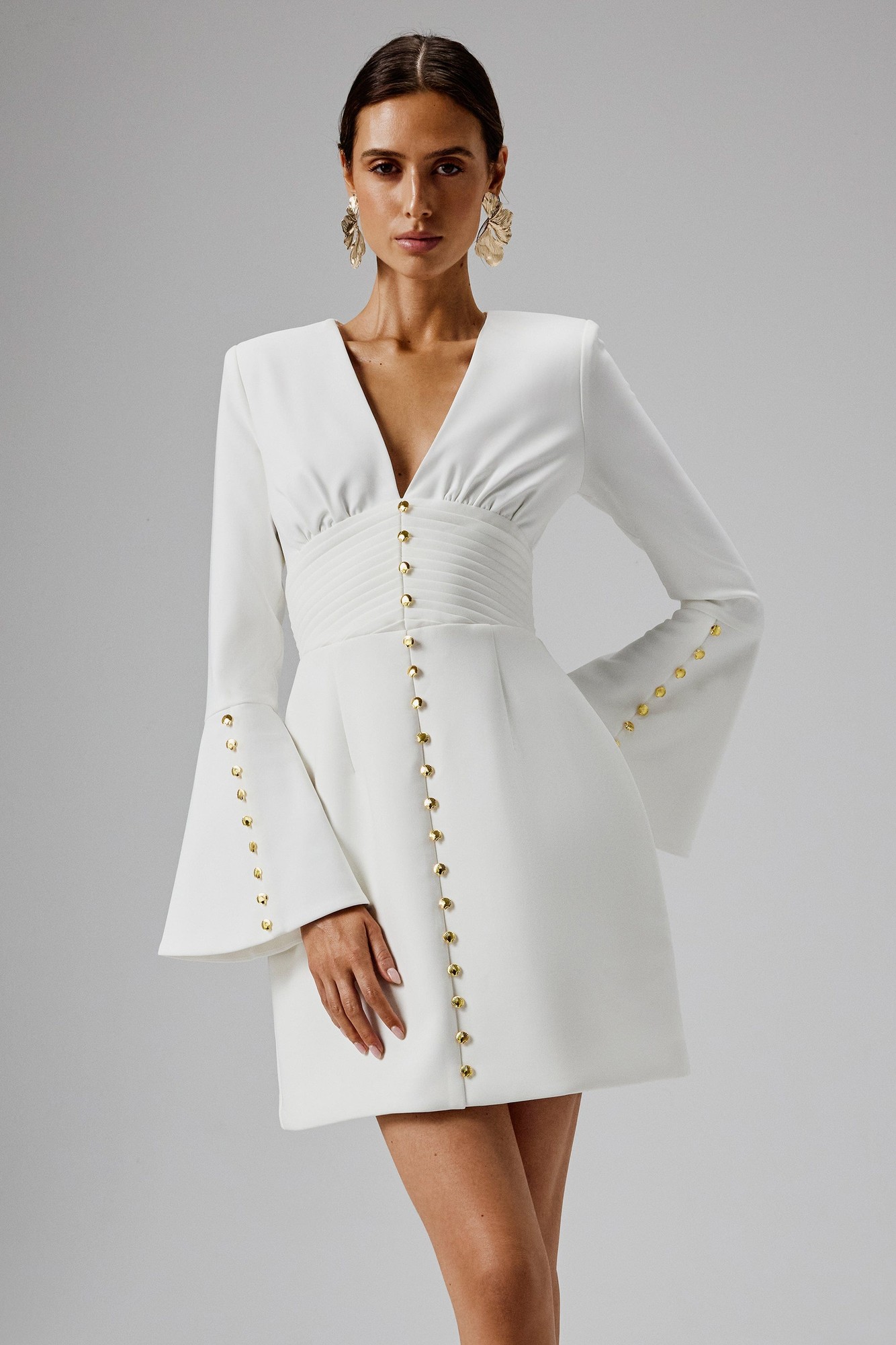 White mini dress with fluted sleeves, deep V-neck, ruched waist, and gold button detailing down the front, made from a smooth fabric, styled for a chic look.