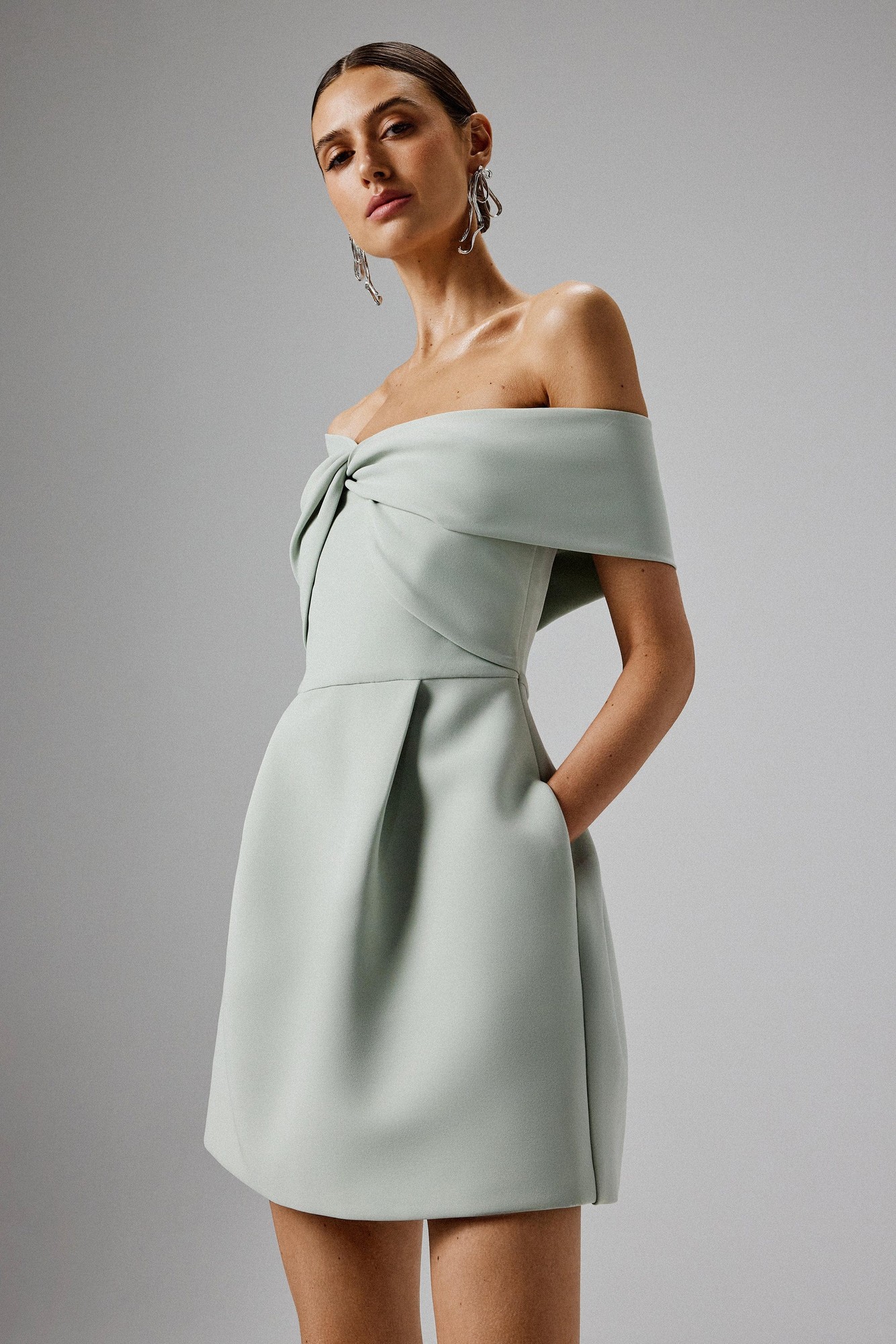 Sage green mini dress with a twisted one-shoulder design, structured bodice, off-shoulder sleeves, and side pockets, made from a smooth material.