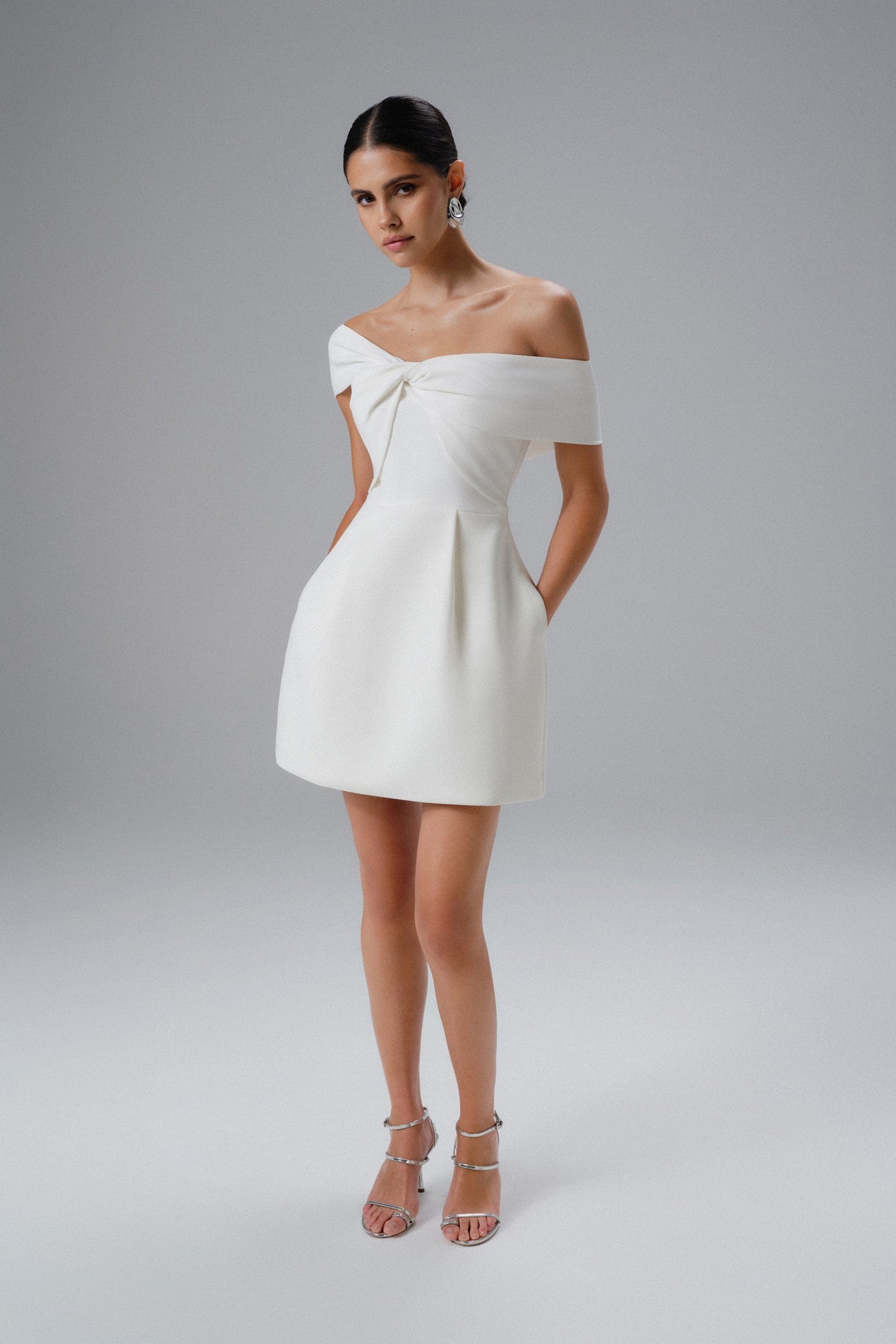 White mini dress featuring a twisted one-shoulder design, structured fabric, and side pockets, styled with strappy silver heels and statement earrings.