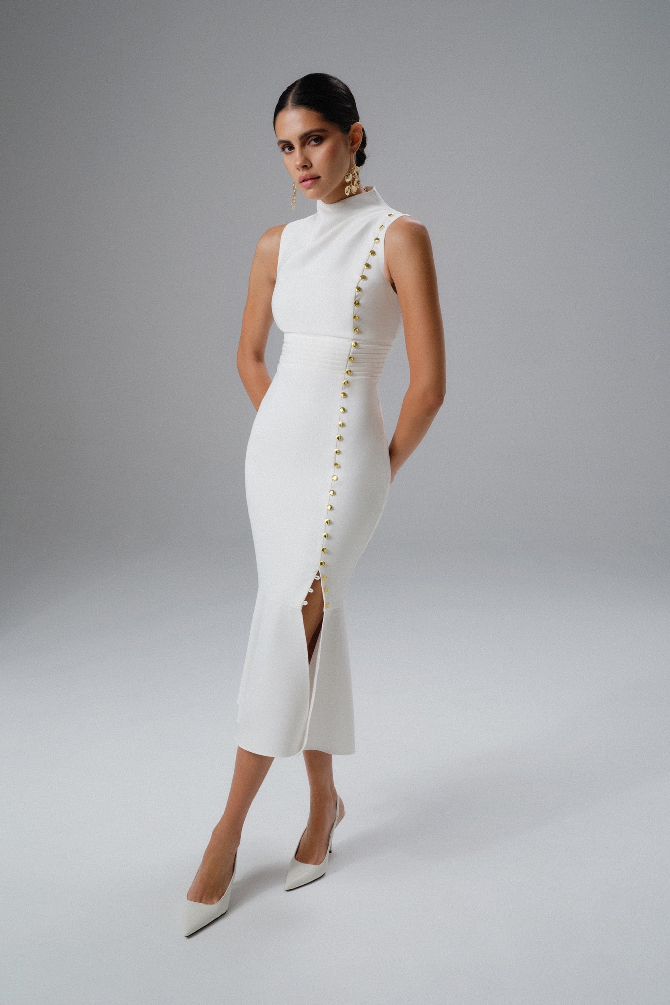 White high neck midi dress with micro shell button detailing along the side, featuring a fitted silhouette and a front slit, styled with pointed toe heels and statement earrings.