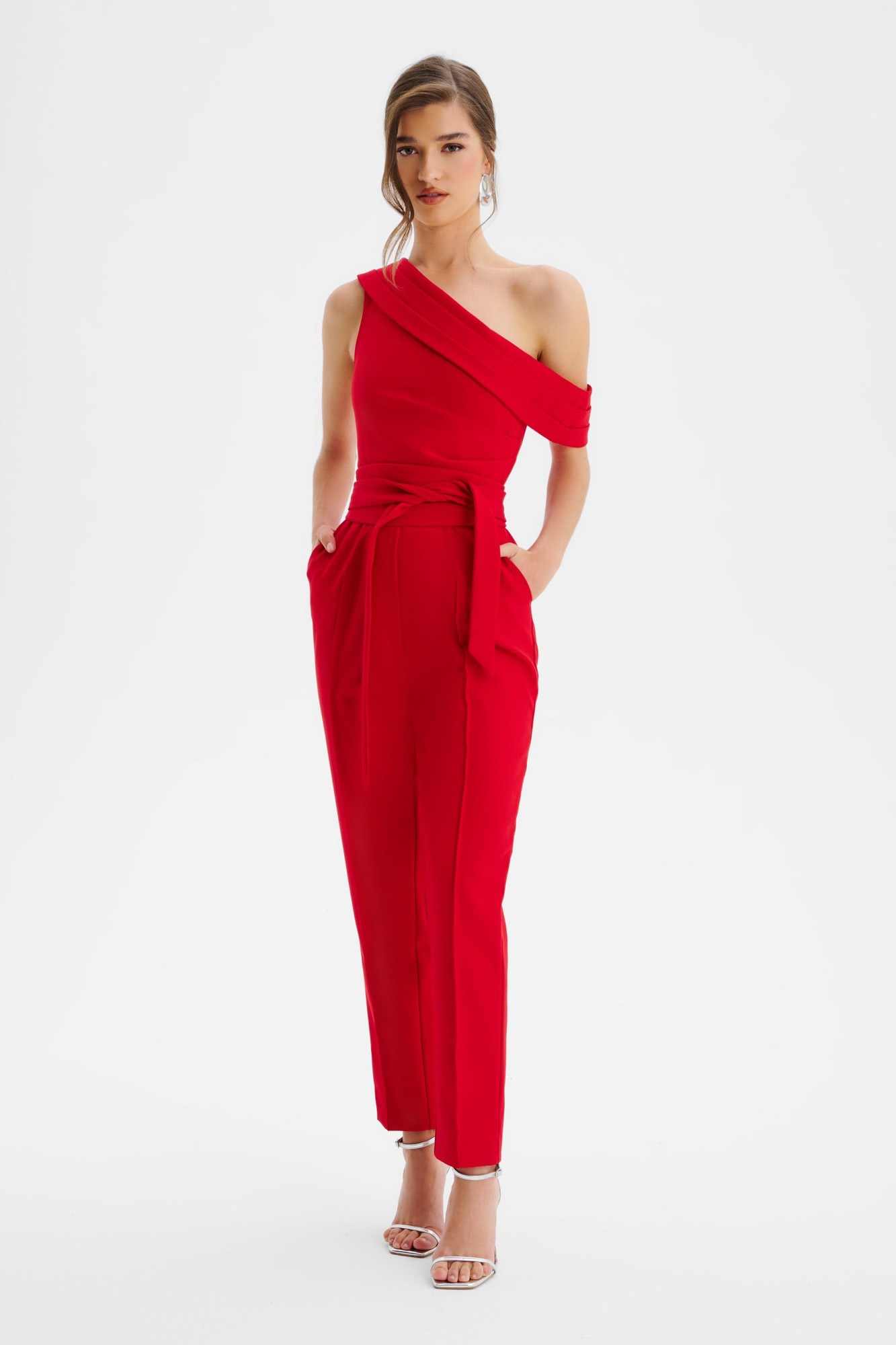 Red off-shoulder jumpsuit featuring pleated detailing, a belted waist, and side pockets, made from a smooth fabric, styled with strappy heels.