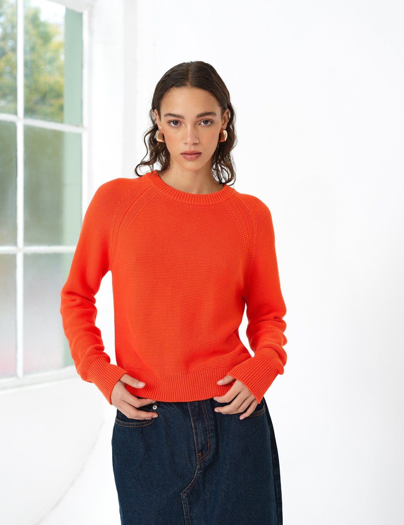 Bright orange knit long sleeve jumper featuring a ribbed crew neckline and raglan sleeves, paired with a dark denim skirt, set against a bright, airy background.