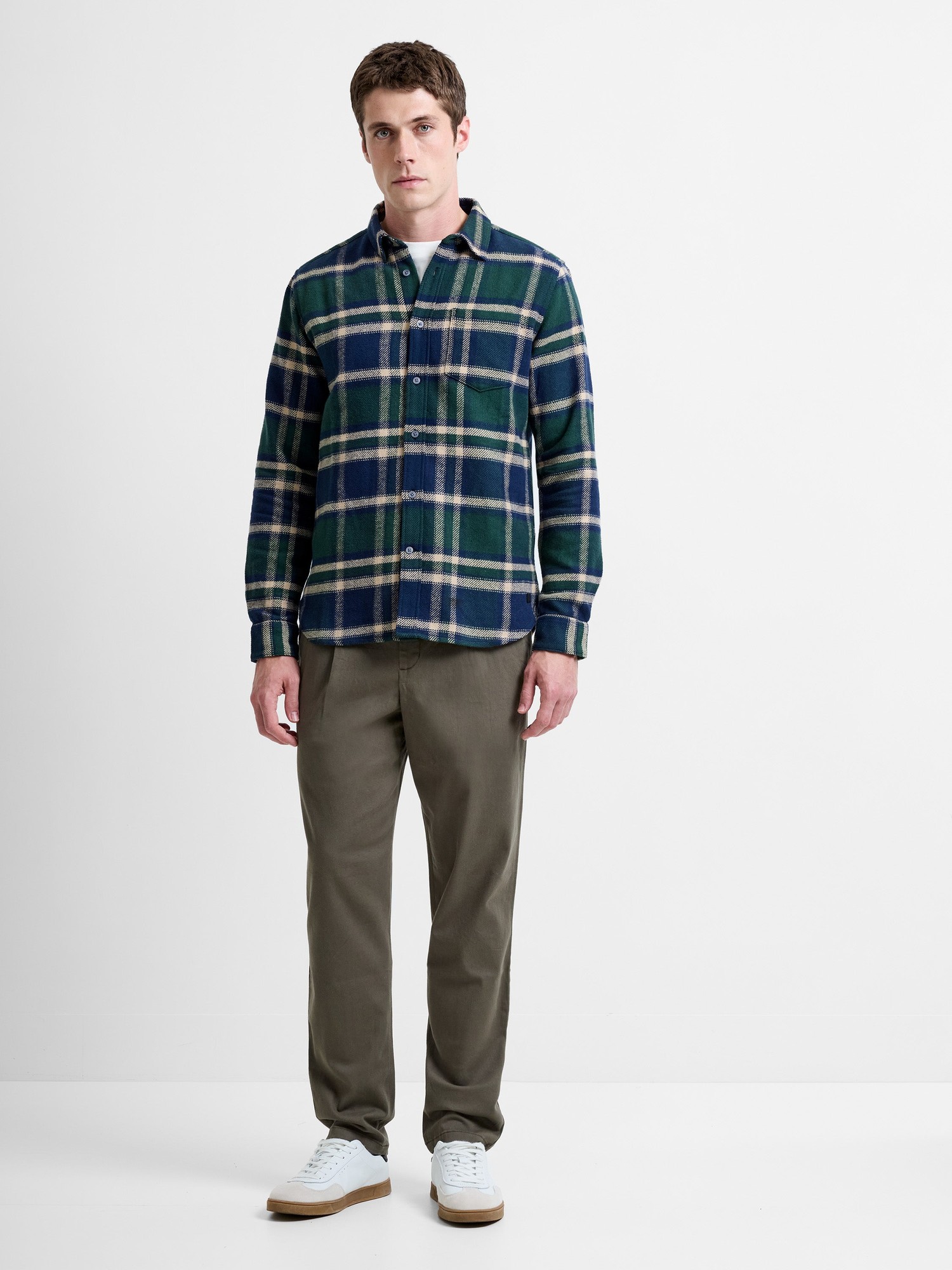 Long sleeve check shirt in navy, green, and cream plaid, featuring a button-down collar, chest pocket, and relaxed fit, paired with olive green trousers and white sneakers.