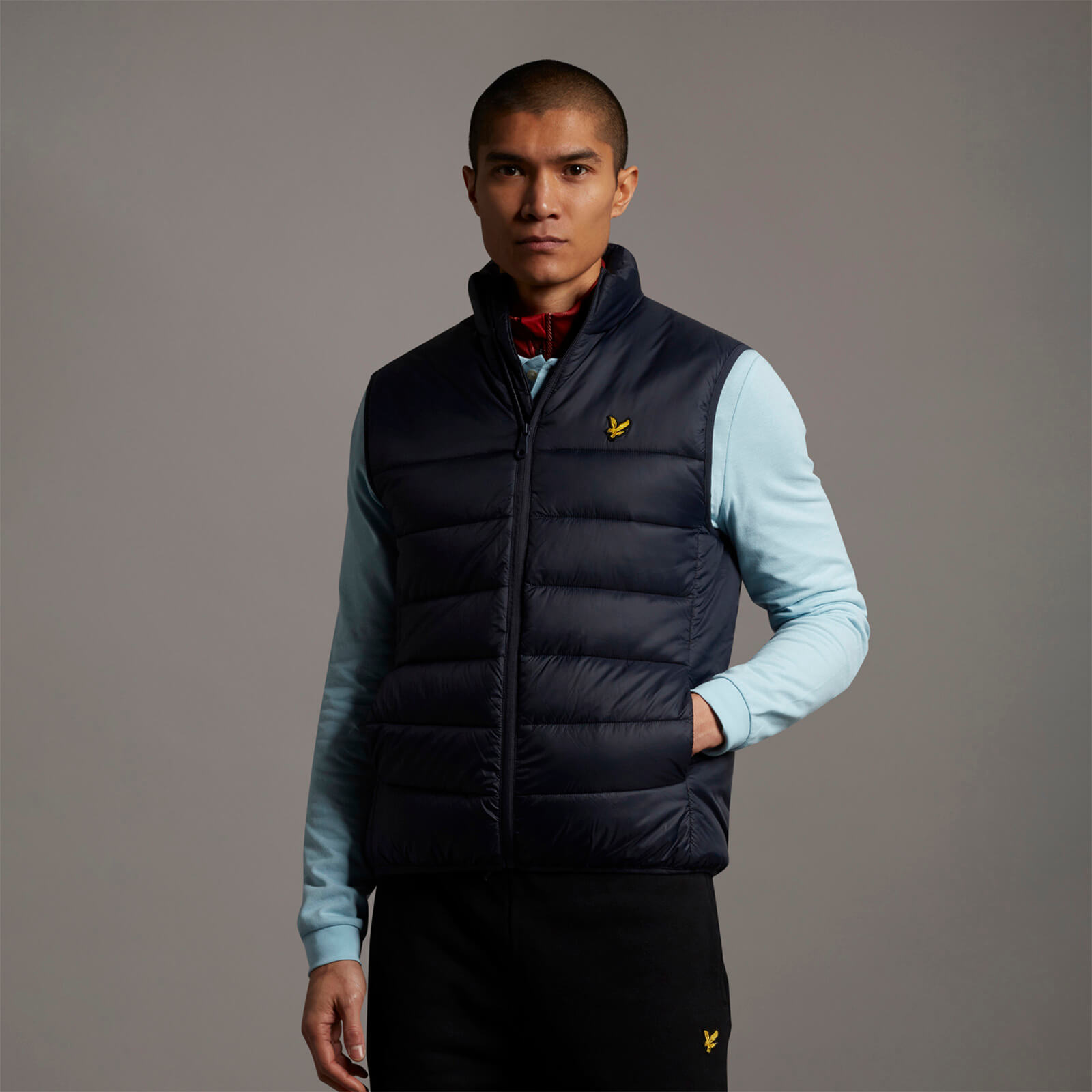 Navy blue puffer jacket with a zip front, featuring a high collar, side pockets, and the Lyle & Scott logo embroidered in yellow on the chest, worn over a light blue long-sleeve shirt.