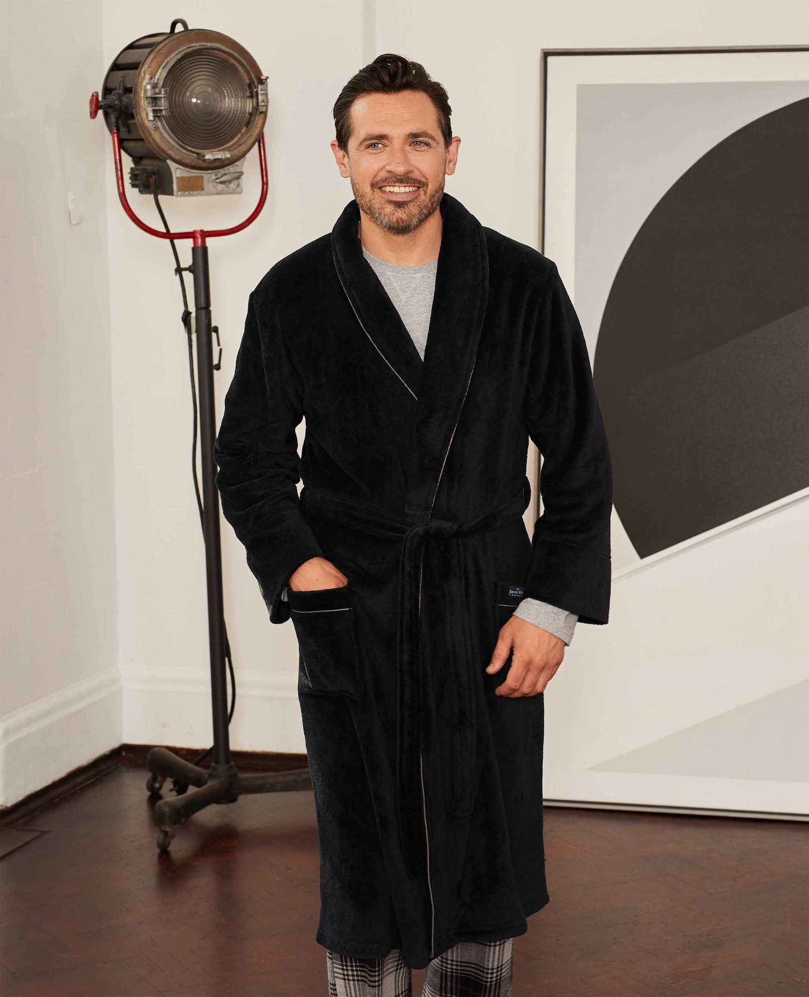 Black supersoft fleece dressing gown with piping, featuring a shawl collar and side pockets, worn by a man standing in a stylish interior setting.