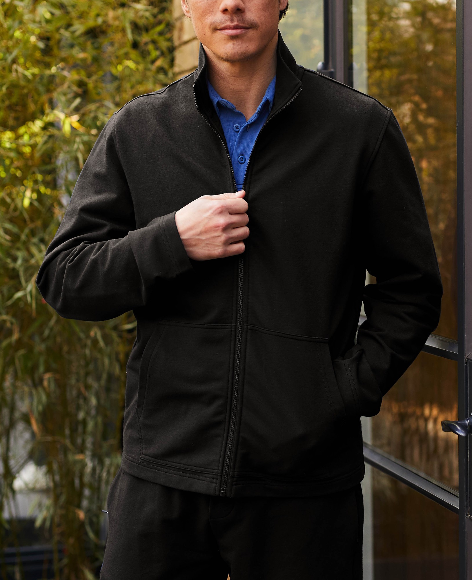 Men's black zip-up sweatshirt made from a stretch cotton blend, featuring a high collar and front pockets, worn over a blue shirt, displayed in a natural outdoor setting.