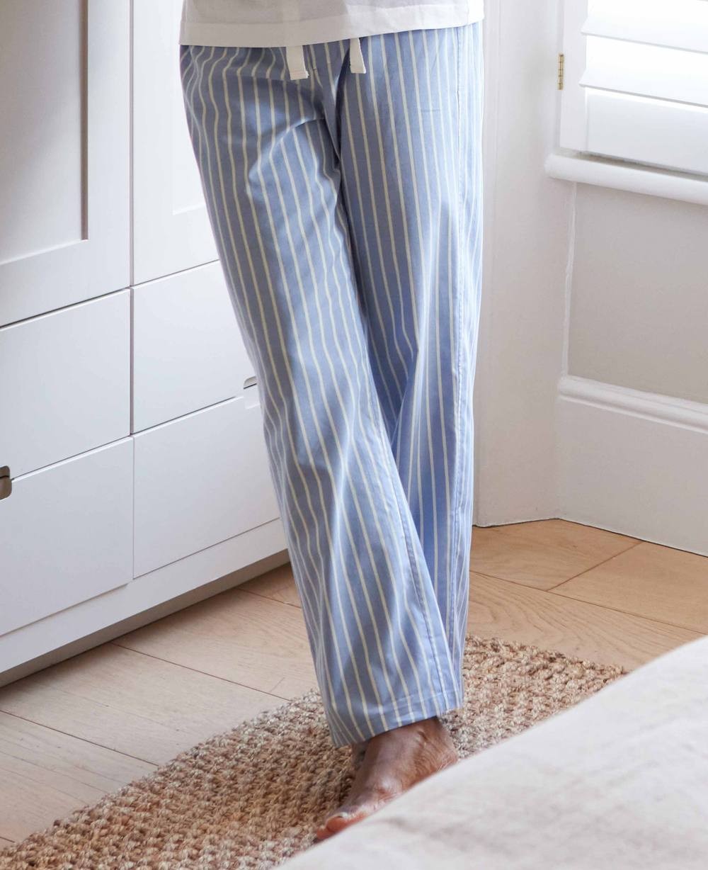 Pale blue cotton twill pyjama bottoms with wide white stripes, featuring an elastic waistband and drawstring, shown on a person standing next to a light-colored rug and furniture.