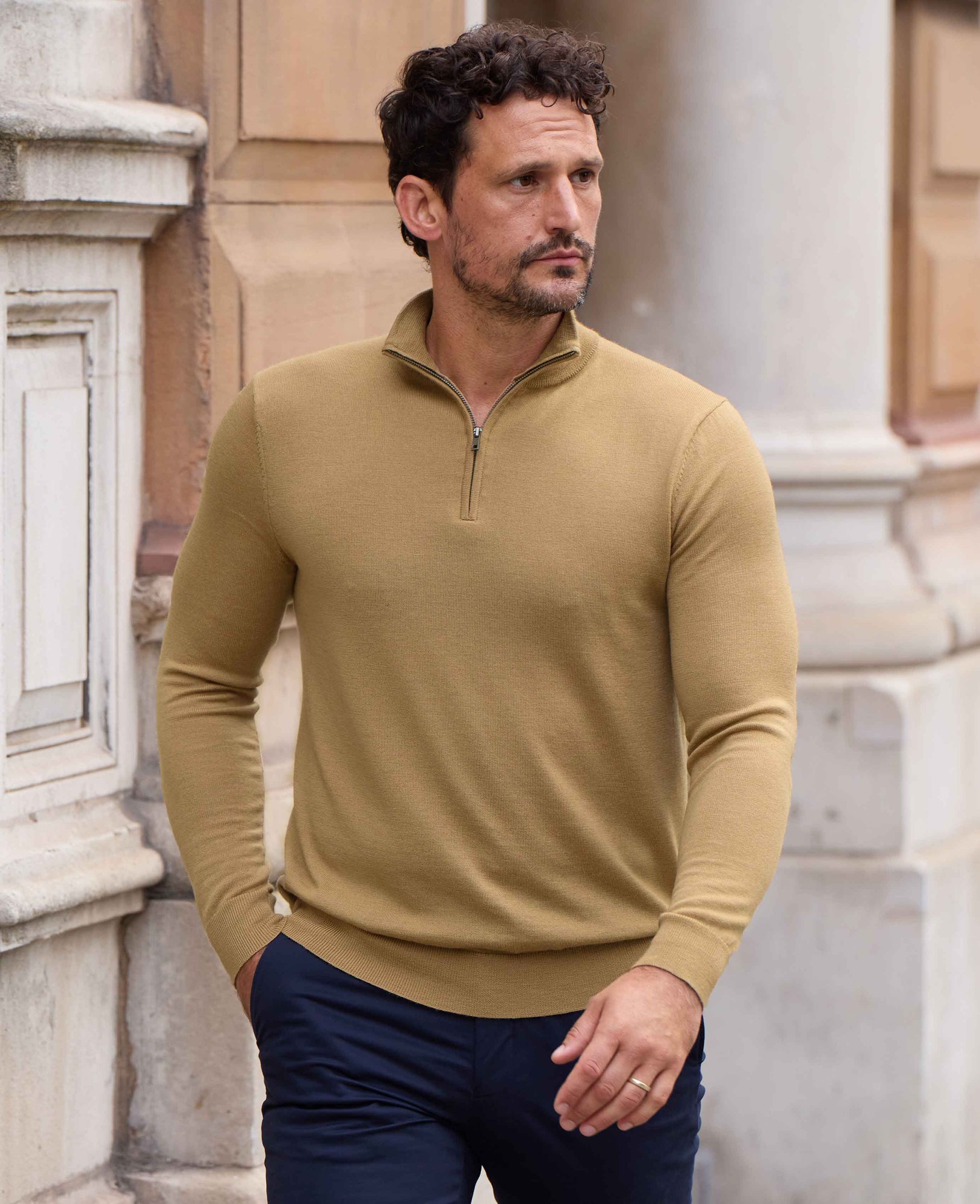 Tan merino wool zip neck jumper for men, featuring a slim fit and ribbed hem, worn by a man with short curly hair, set against a classic architectural background.
