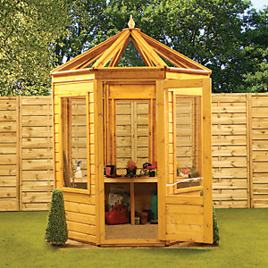 Octagonal wooden greenhouse in natural finish, featuring a sloped roof, large glass panels, double doors, and a wooden shelf inside, set against a green lawn and wooden fence.