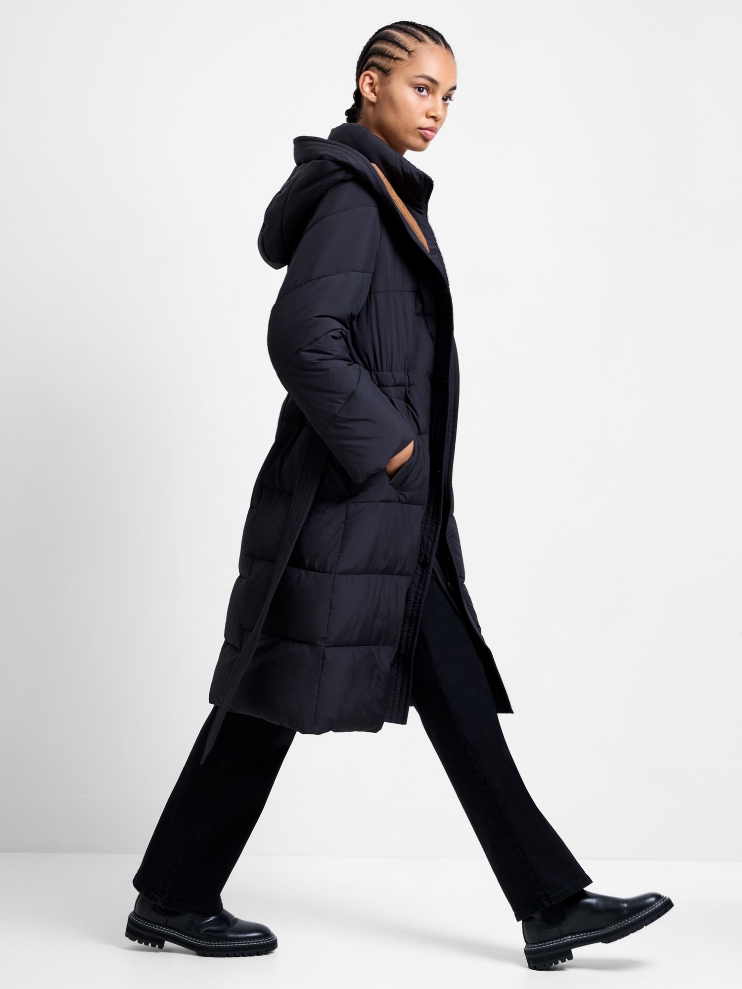 Long black belted hooded coat with a quilted design, featuring a relaxed fit, side pockets, and a stylish silhouette, paired with black pants and chunky black boots.