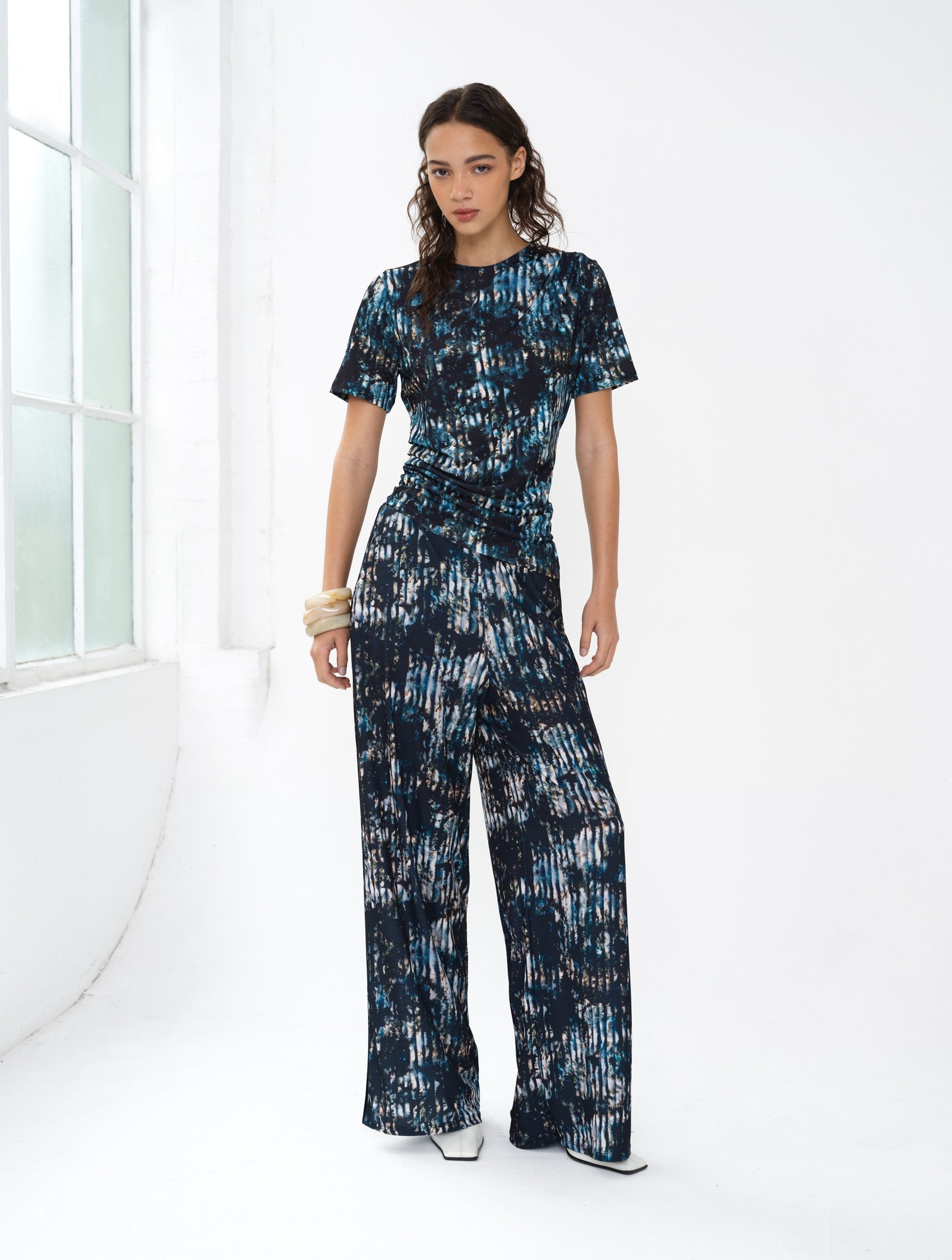 Black multi-colored draped top featuring a tie-dye pattern, short sleeves, and a fitted silhouette, paired with wide-leg pants for a stylish, modern look.