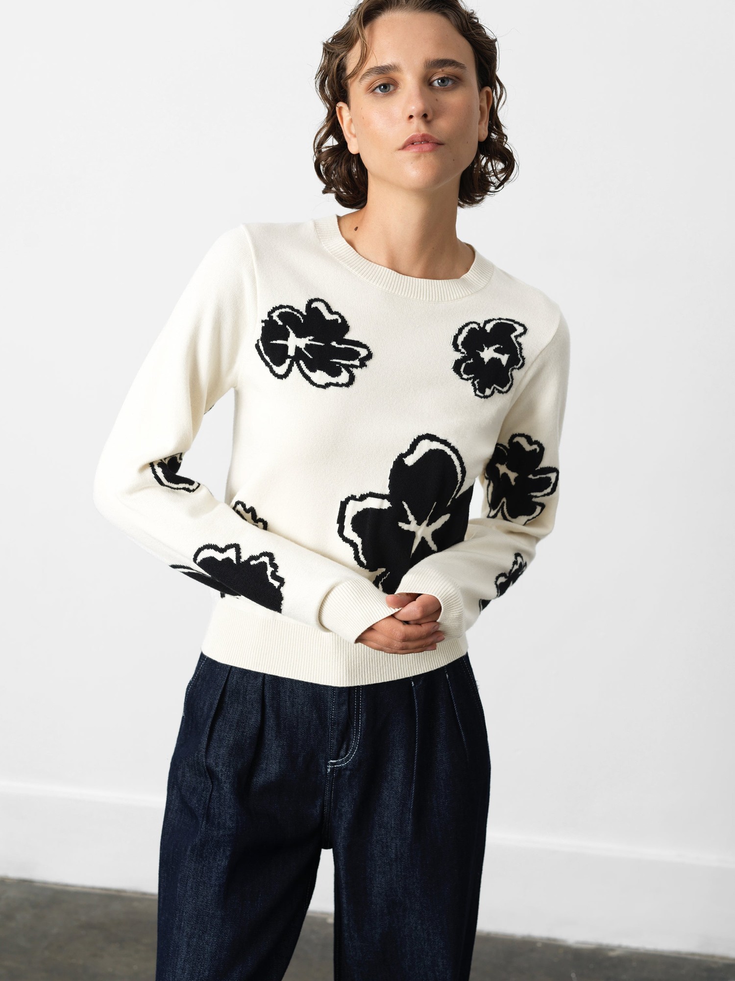 White floral jacquard jumper featuring black floral patterns, round neckline, and ribbed cuffs, styled with dark denim trousers.